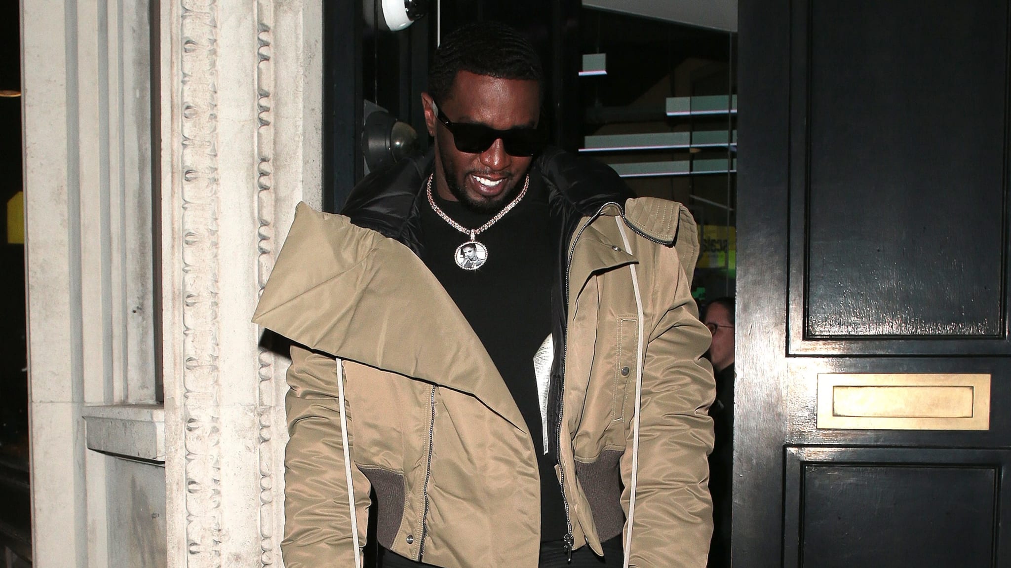 Sean "Diddy" Combs wearing sunglasses and a beige jacket, smiling as he exits a building.