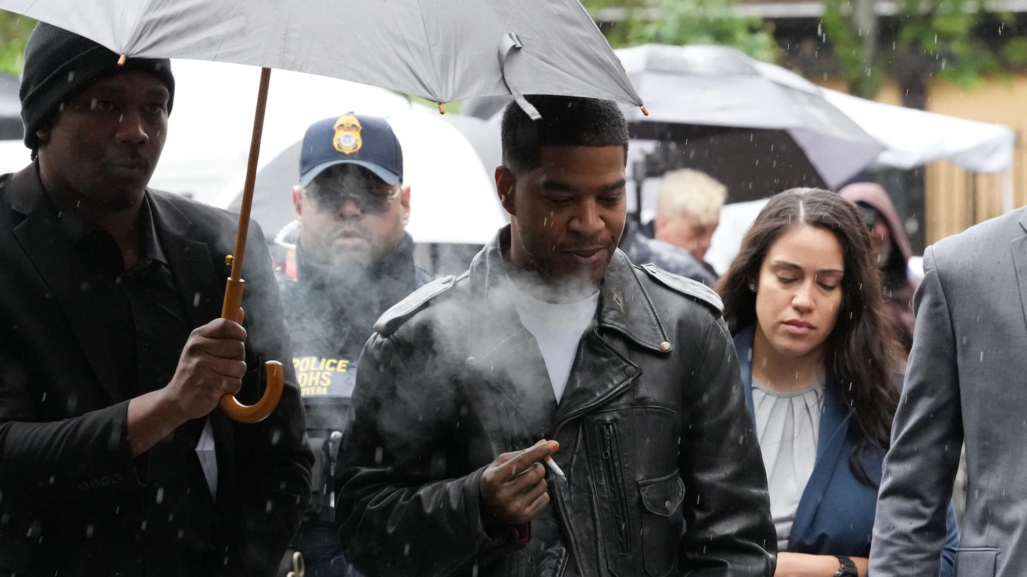 A group of people walking in the rain, one man holding an umbrella. A man in a leather jacket and a woman in a suit are visible.