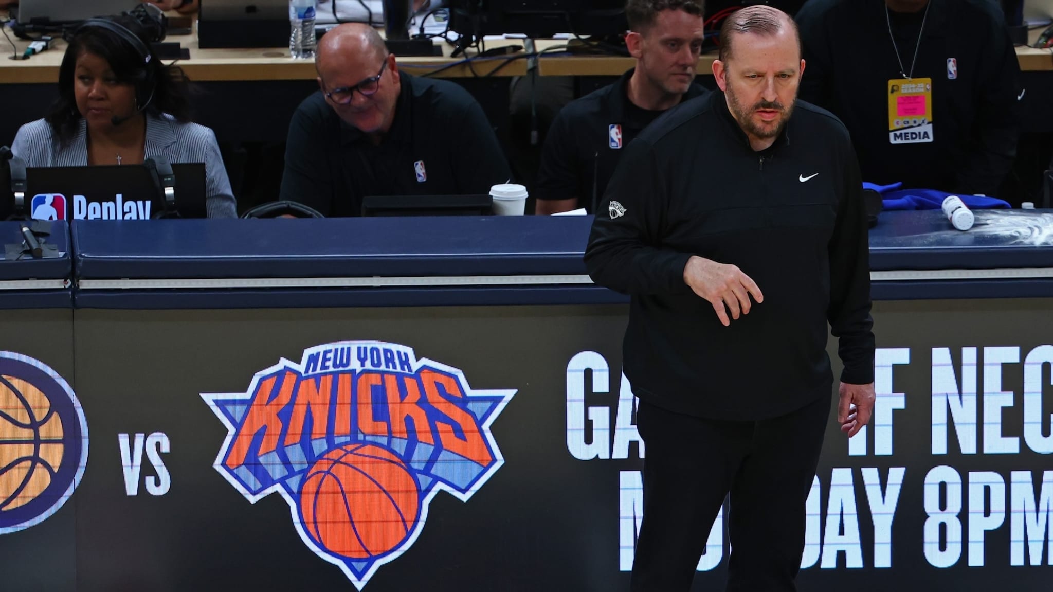 A basketball coach stands near the New York Knicks logo, with media personnel seated behind him at a sports event.