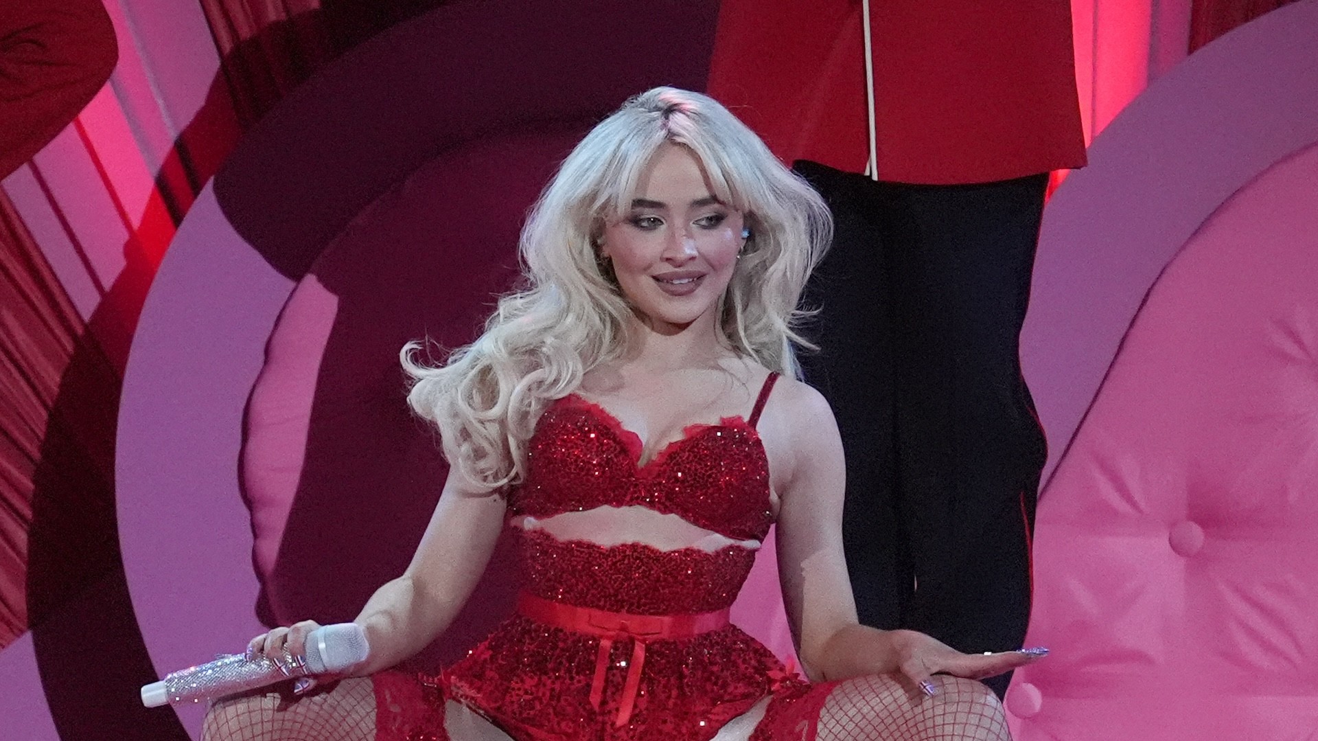 Sabrina Carpenter in a red sparkly outfit and fishnet stockings is on stage, holding a microphone, with a pink and red backdrop.