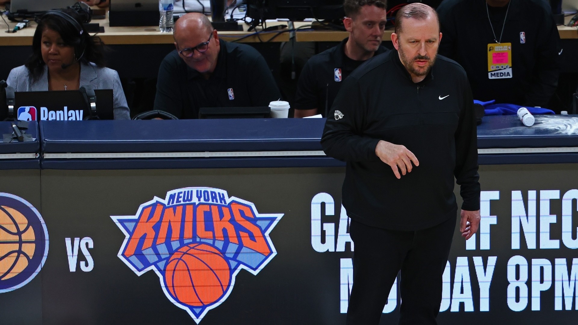 A basketball coach stands near the New York Knicks logo, with media personnel seated behind him at a sports event.