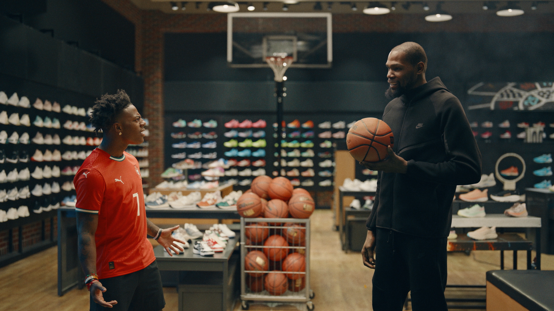 IShowSpeed in a red Puma jersey talks to Kevin Durant in a black hoodie holding a basketball in a sneaker store with basketballs and shoes.