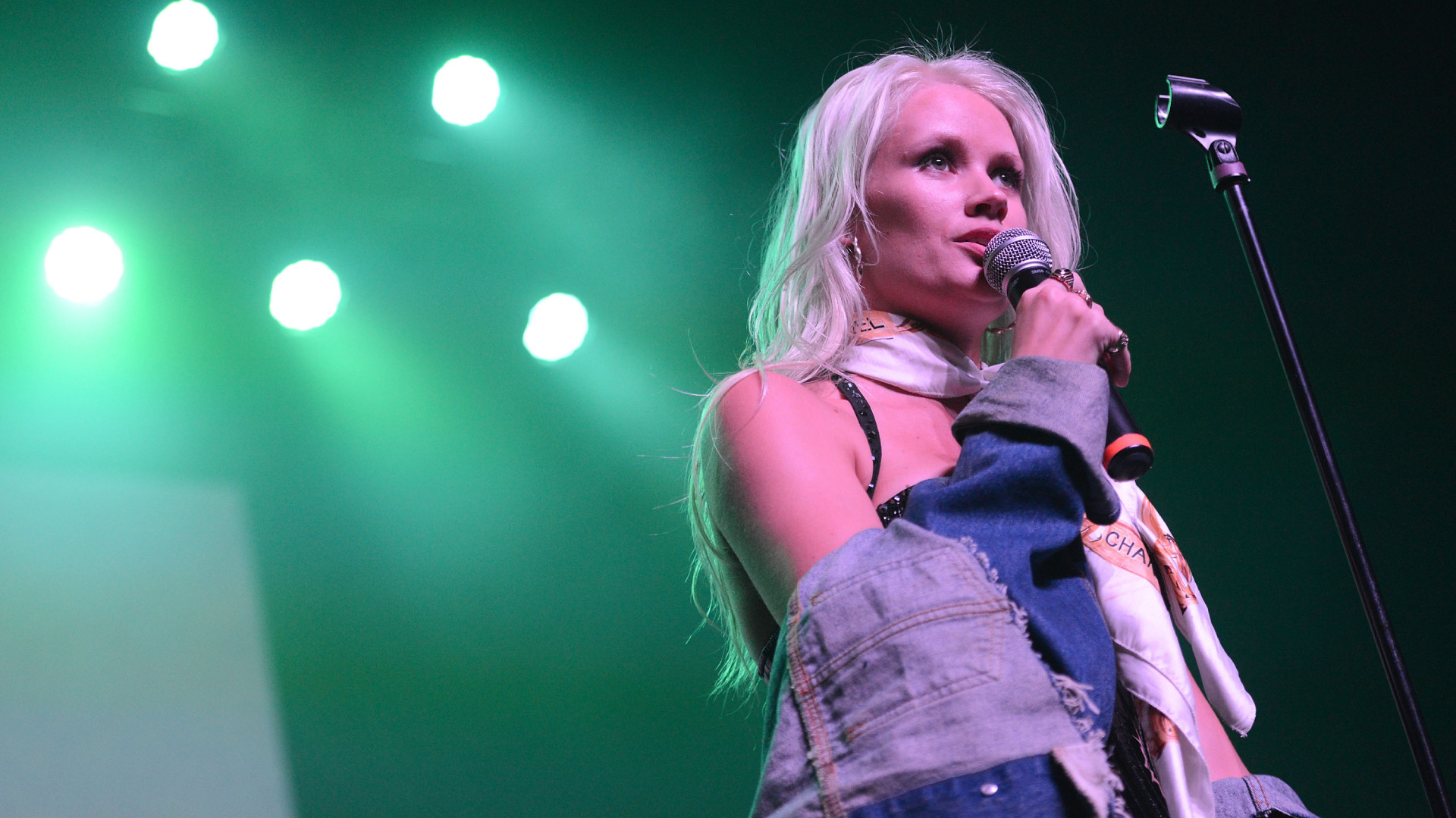 Singer Lauren Bennett with long blonde hair sings into a microphone on stage, wearing a denim jacket and scarf, under green stage lights.