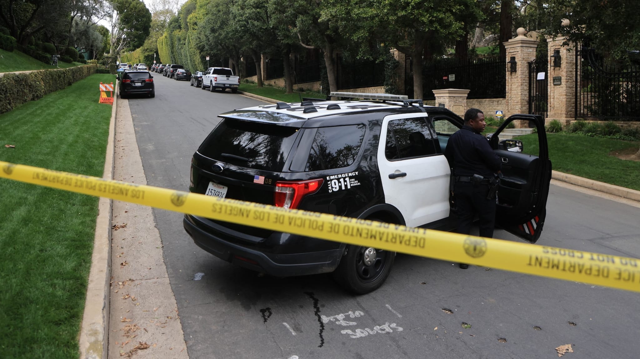 A police SUV is parked on a residential street, with an officer standing beside it. Yellow police tape blocks the road.
