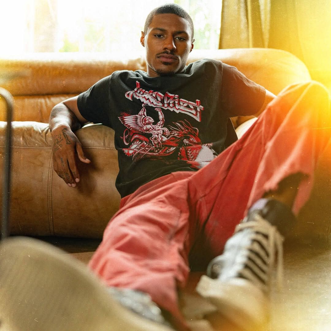 Man lounging on a couch wearing a black graphic T-shirt, red pants, and sneakers, with a relaxed expression.