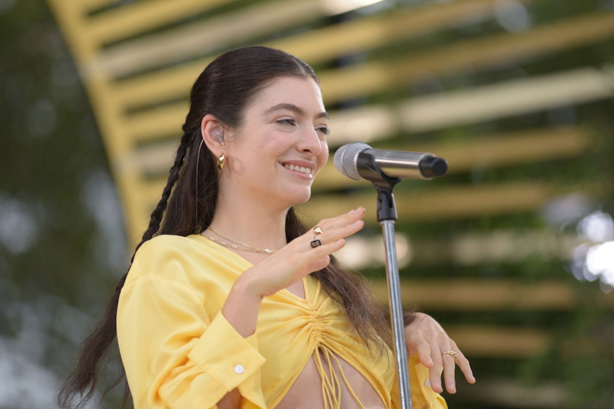 Lorde performing on stage, wearing a yellow outfit, smiling, with a microphone in front of her.
