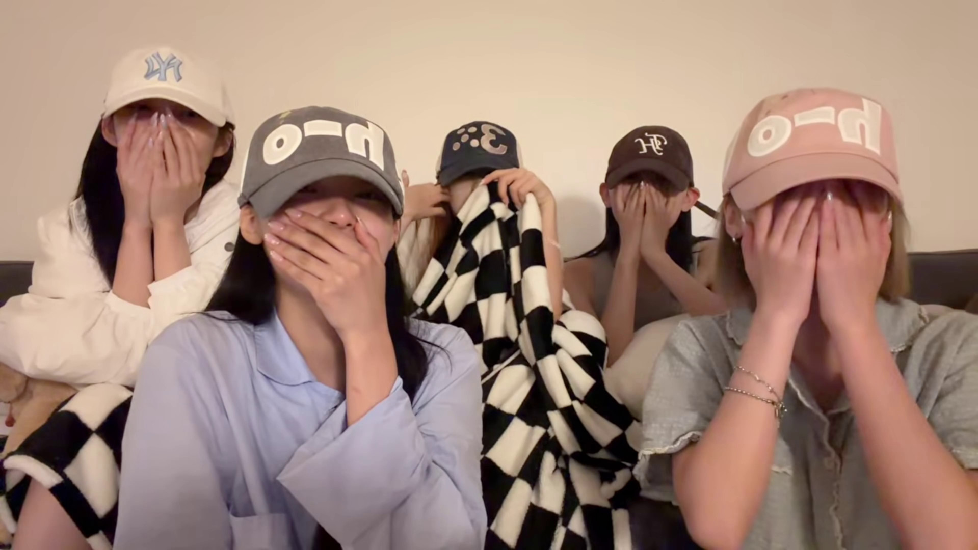 Five members of the K-pop group RESCENE sitting on a couch, wearing caps and covering their faces with their hands.