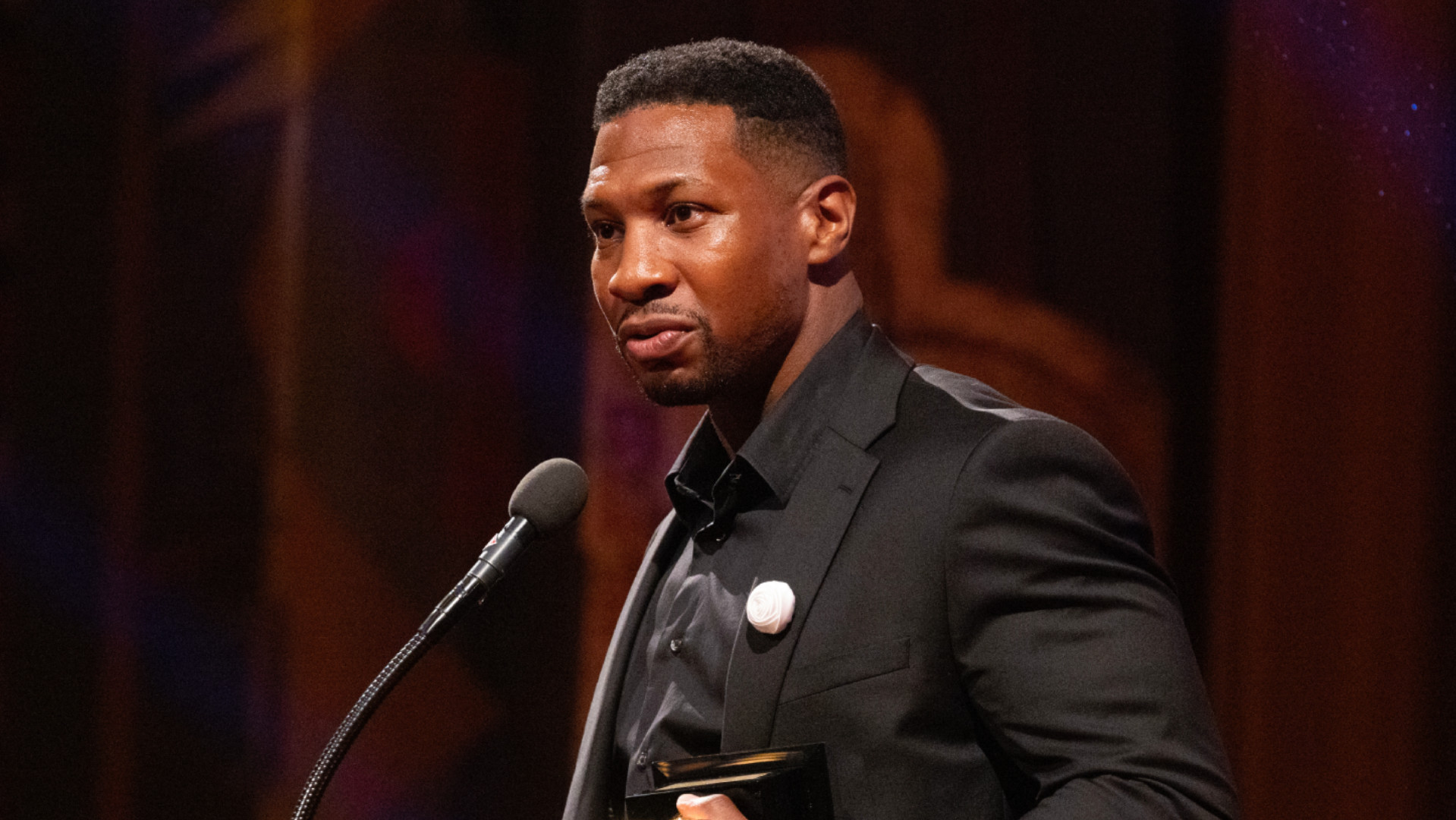 Jonathan Majors in a black suit stands at a microphone holding an award, speaking at an event.