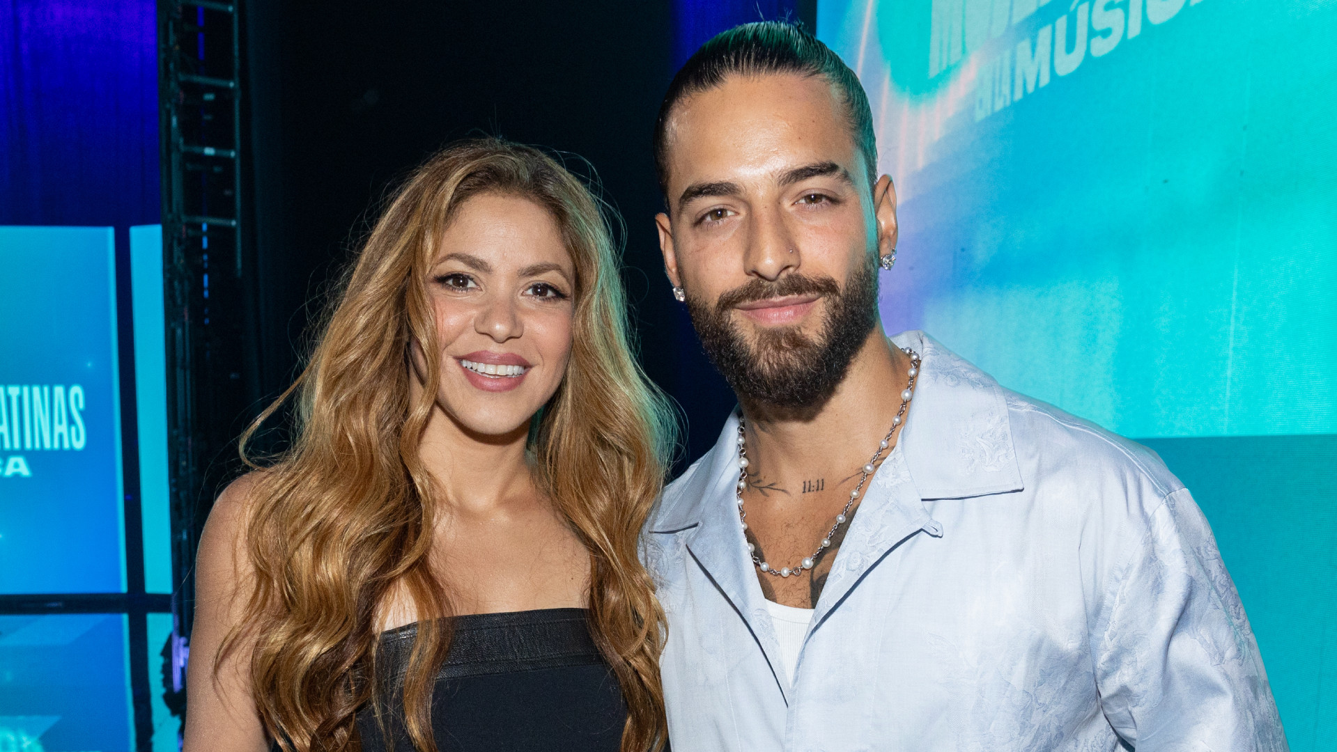 Shakira and Maluma smiling together at an event, with Shakira in a black dress and Maluma in a light shirt.