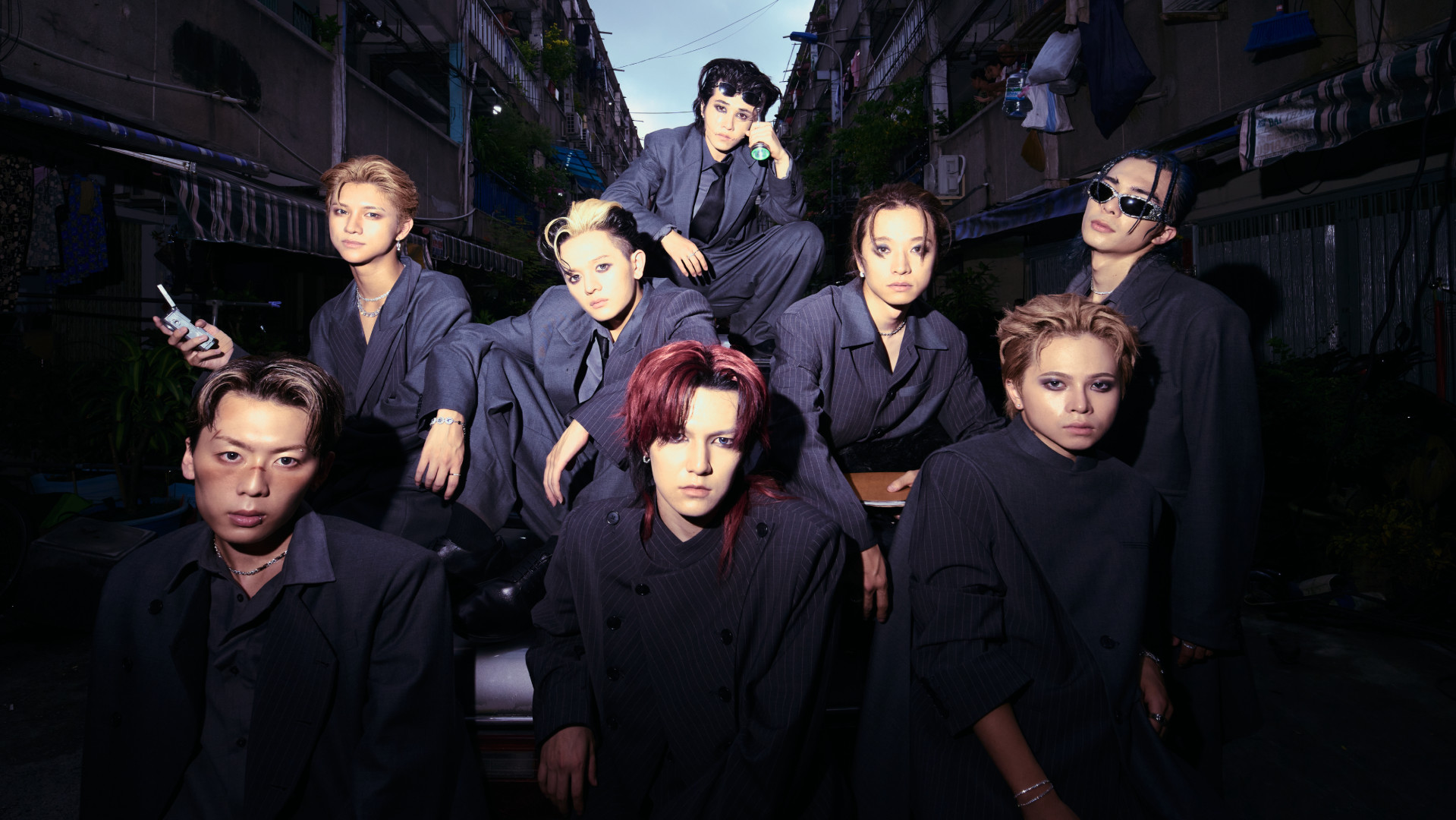 The members of J-pop group ONE OR EIGHT in dark suits pose confidently in an urban alleyway. The scene has a moody, stylish atmosphere.