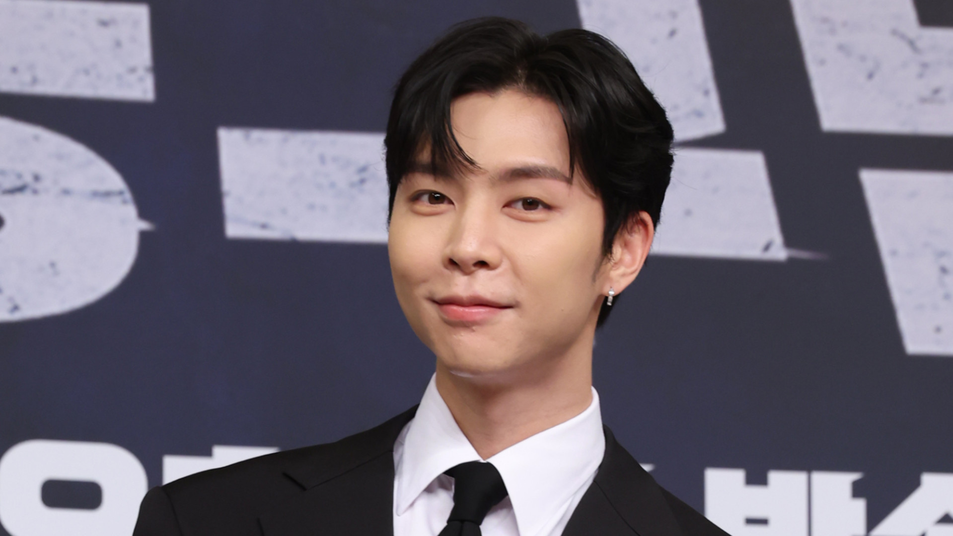 Johnny of NCT 127 in a suit and tie, smiling at an event, standing against a backdrop with text.