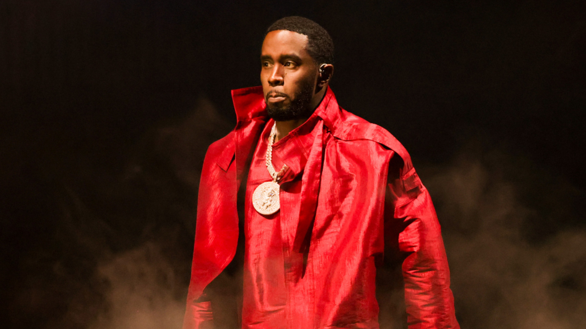 Diddy wearing a red coat and a large pendant necklace, walking through a smoky atmosphere.