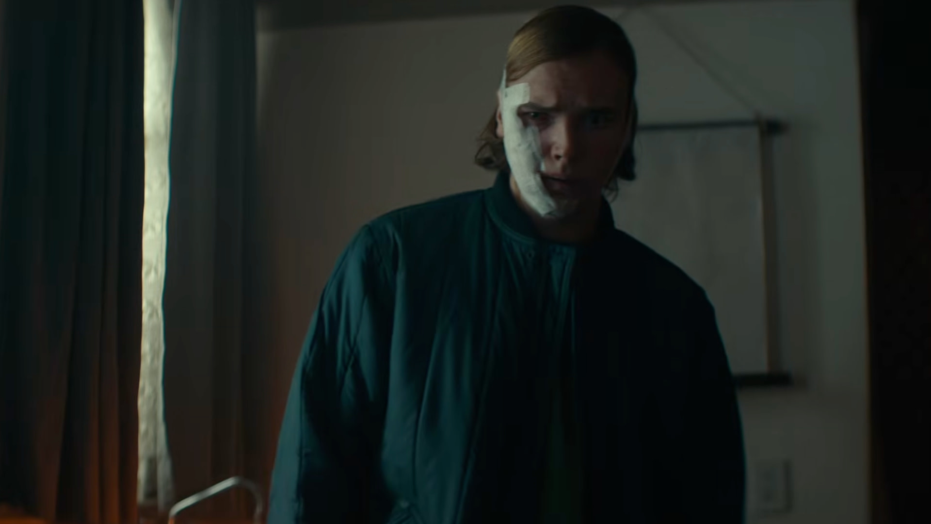 A person with a serious expression, wearing a dark jacket, stands in a dimly lit room with curtains and a wall hanging.
