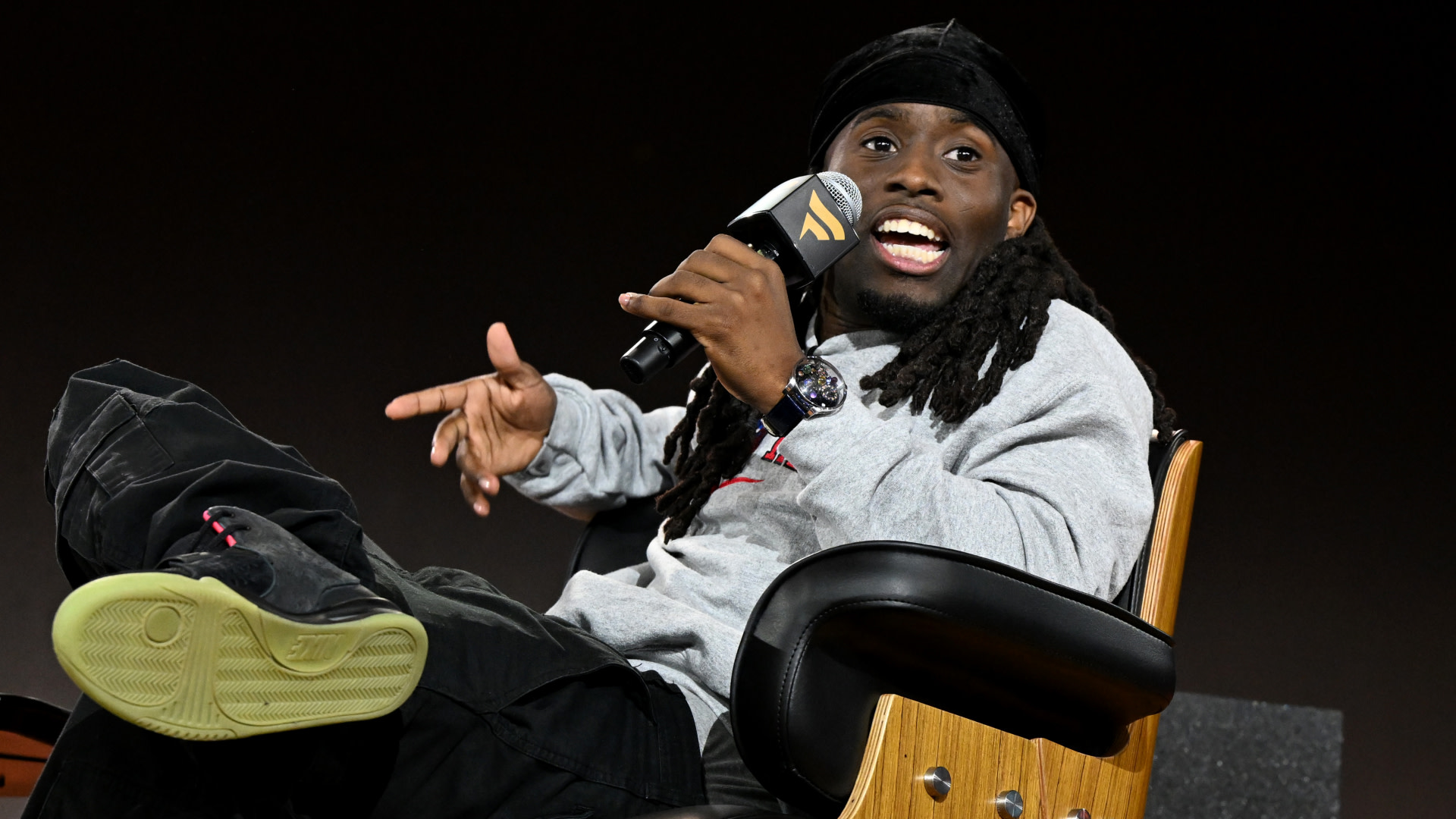 Kai Cenat with long hair and a cap, wearing a gray sweatshirt and black pants, sits in a chair speaking into a microphone.