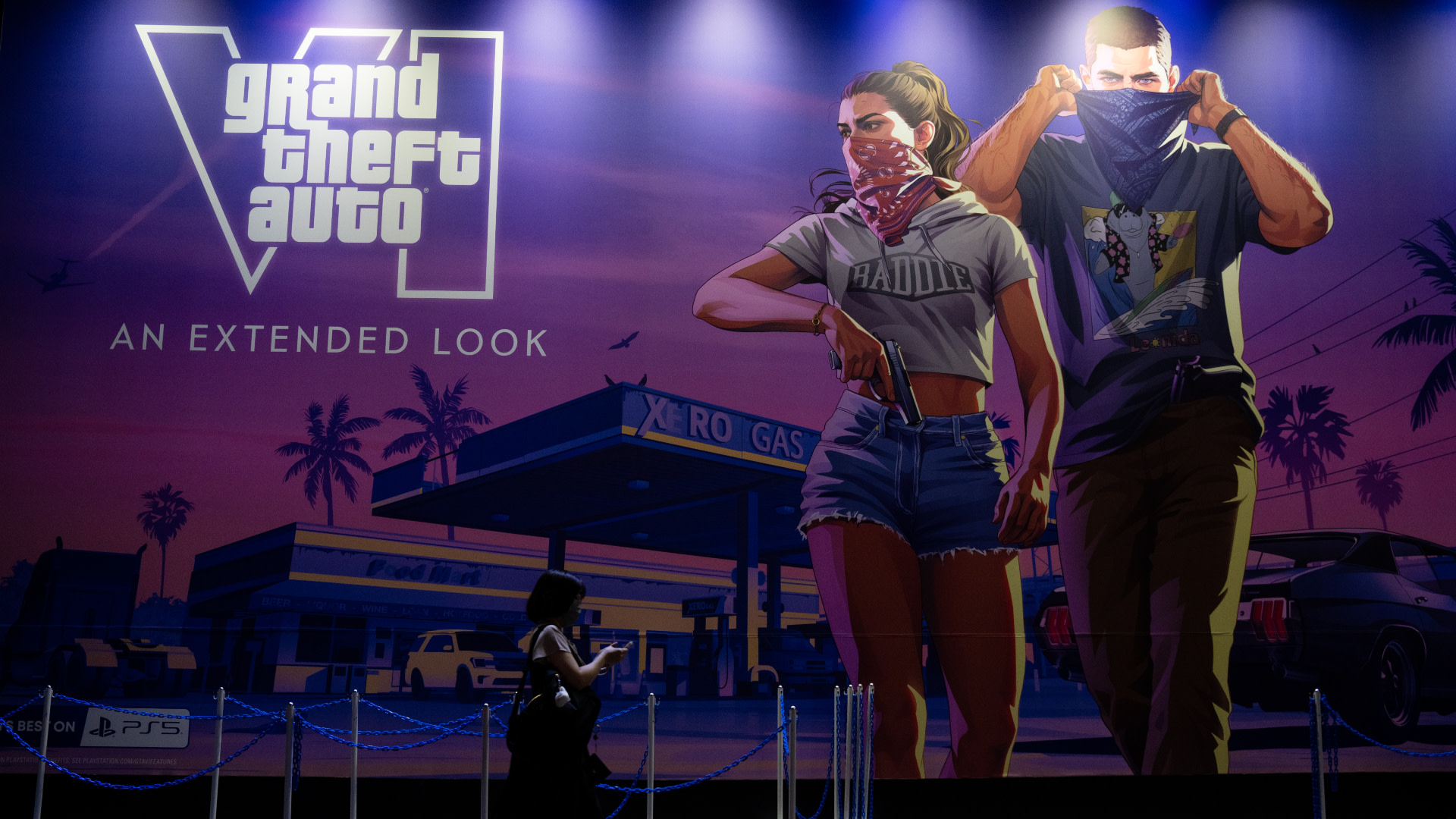 A large "Grand Theft Auto VI" poster with two characters wearing bandanas, standing in front of a gas station. A person is silhouetted below.