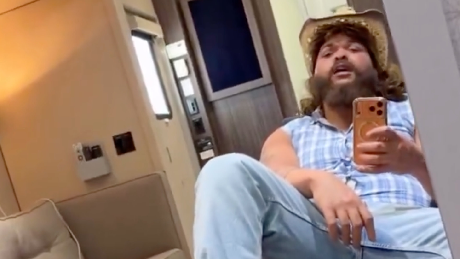 Druski in costume with a beard and cowboy hat takes a selfie in a mirror, wearing a sleeveless plaid shirt and jeans, in a room with a couch.