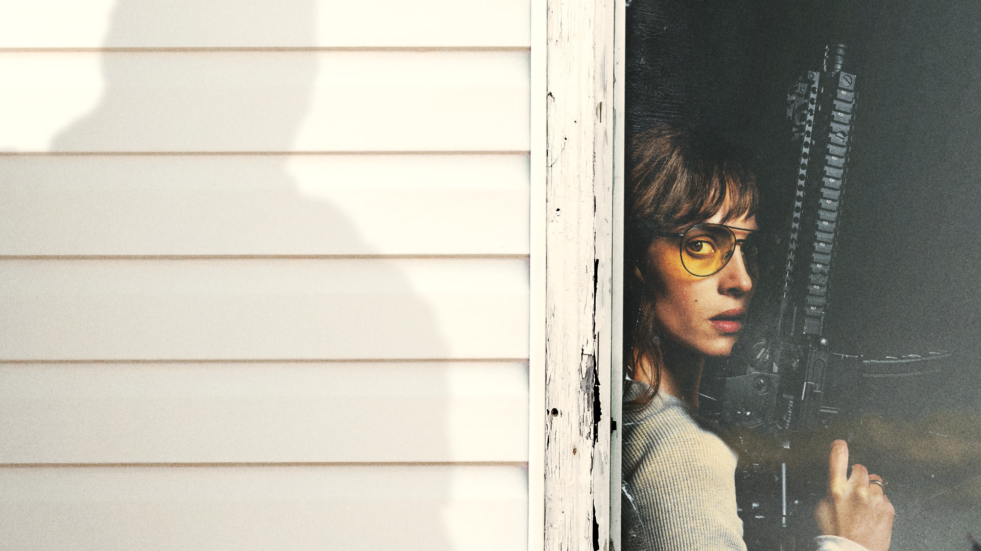 A person with glasses holding a rifle peers out from behind a wall. A shadow is cast on the adjacent siding.