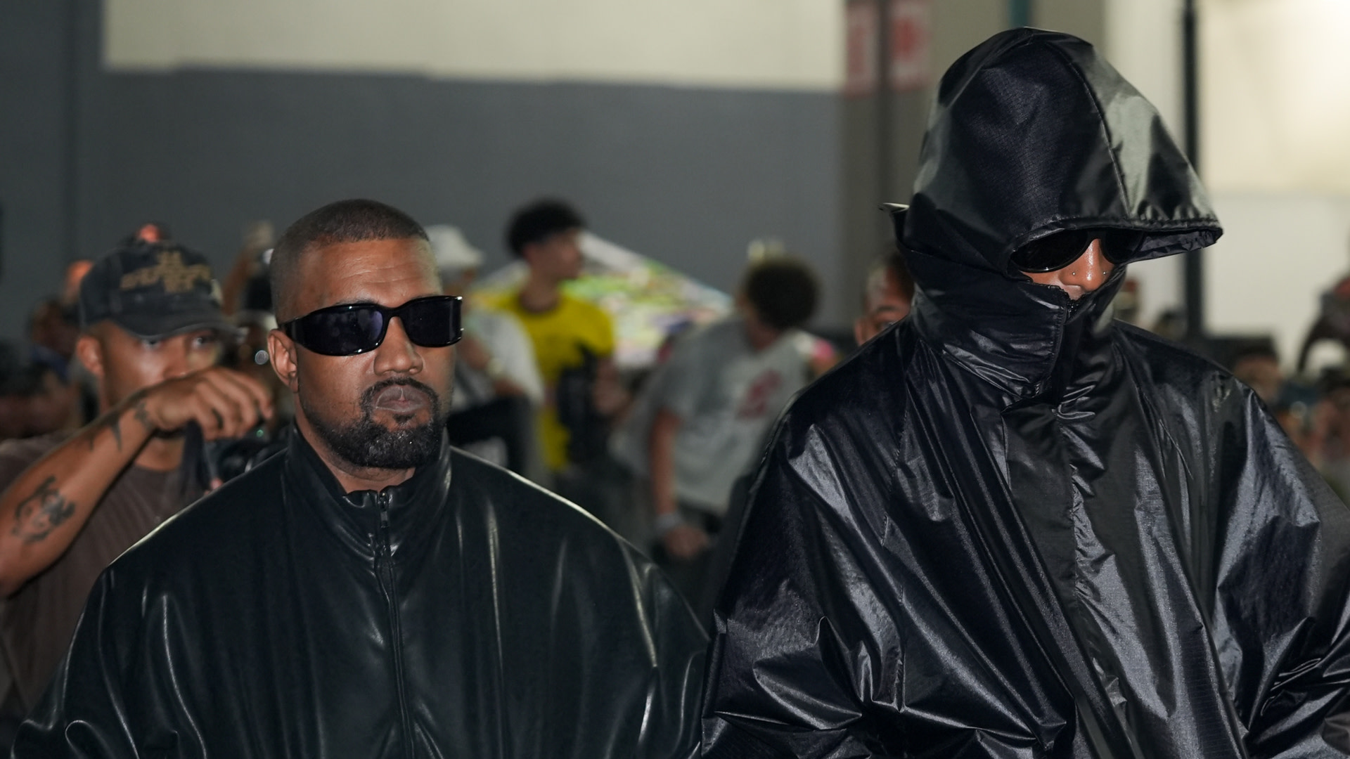 Kanye West in a black leather jacket and sunglasses, walking beside Playboi Carti in a hooded black outfit, surrounded by people.