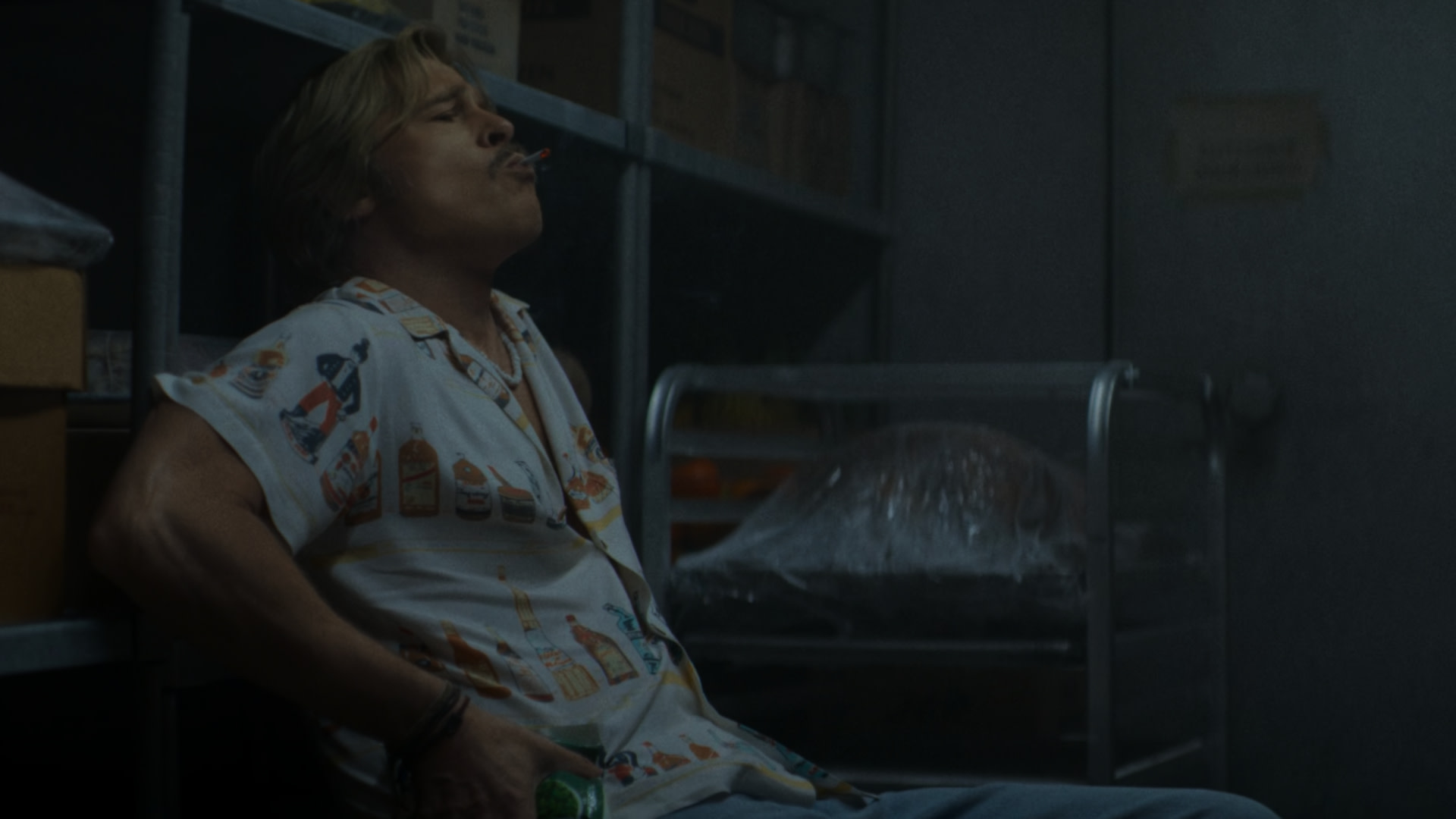 Brad Pitt in a dimly lit room is sitting on a chair, wearing a patterned shirt, holding a drink, and smoking.