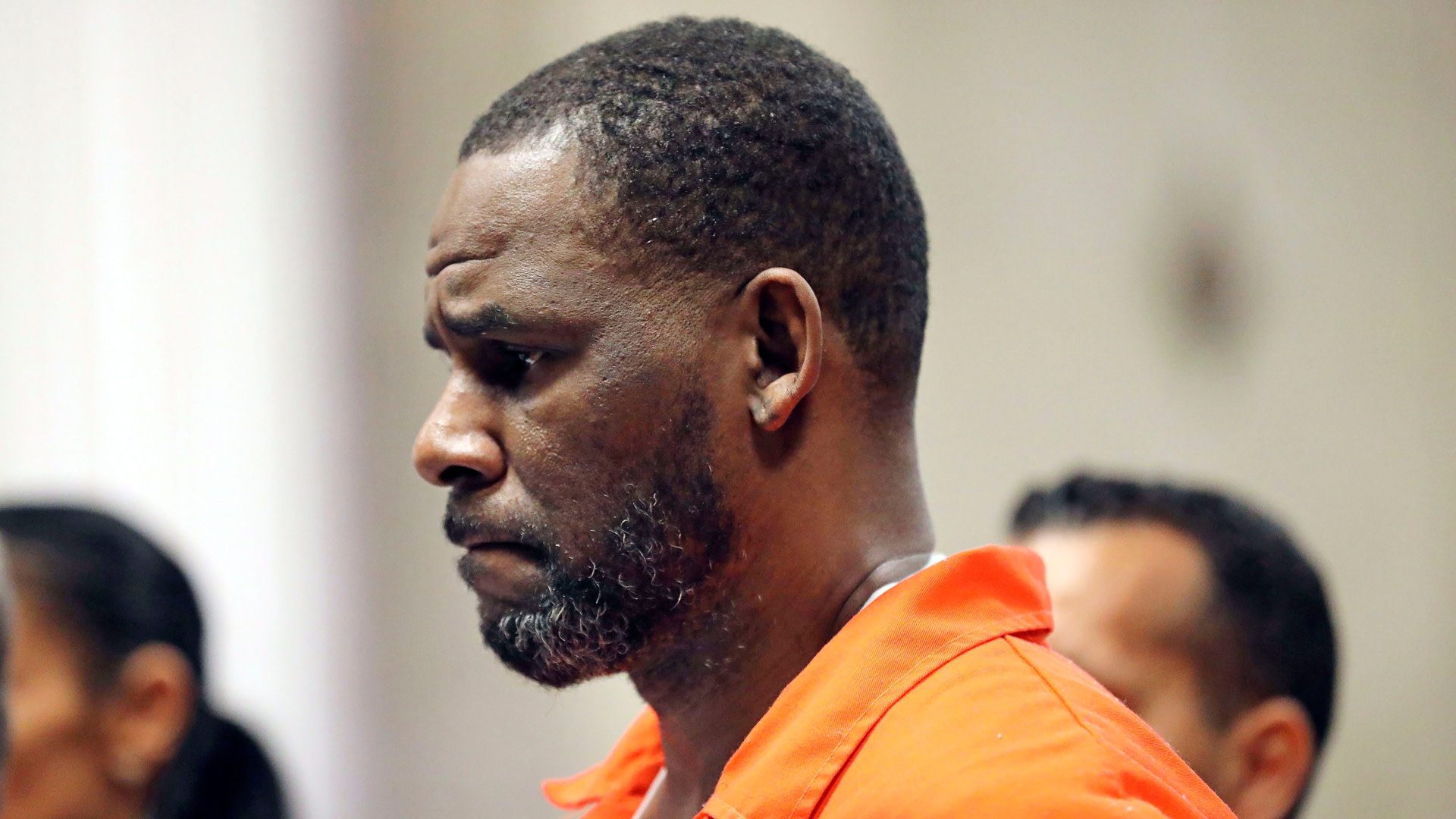 R. Kelly in an orange jumpsuit appears in court, looking down with a serious expression.