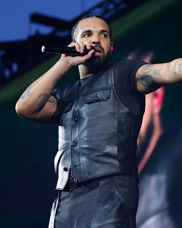 Drake performing on stage, wearing a black sleeveless leather vest, holding a microphone, and pointing towards the audience.