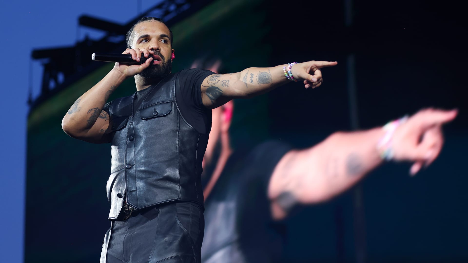 Drake performing on stage, wearing a black sleeveless leather vest, holding a microphone, and pointing towards the audience.
