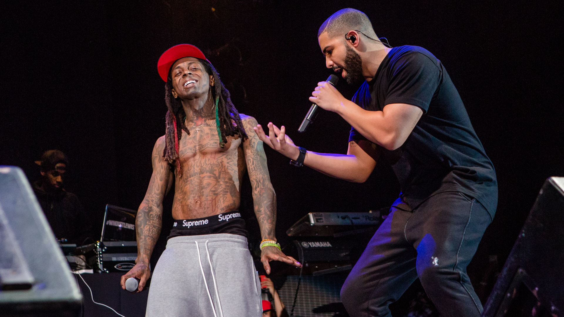 Lil Wayne and Drake performing on stage; Lil Wayne is shirtless with a red cap, and Drake is holding a microphone, wearing black.