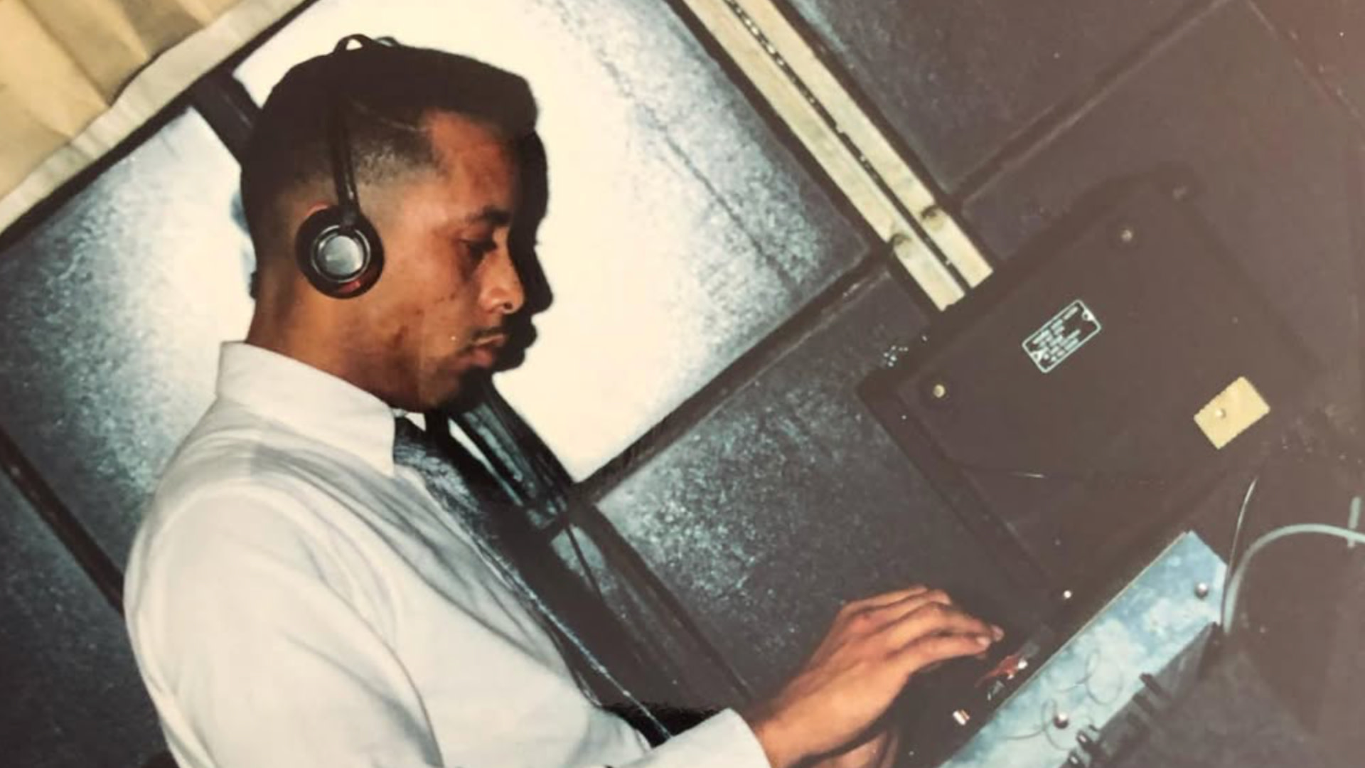 Tone Capone wearing headphones and a white shirt, focused on DJ equipment, set against a backdrop of large speakers.