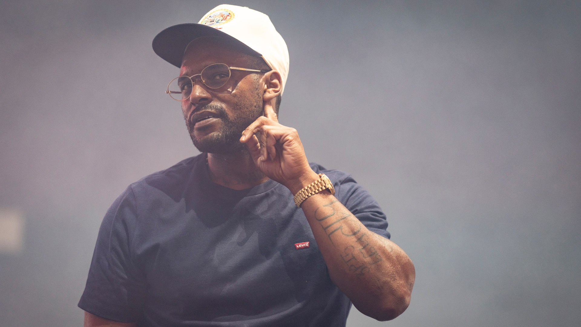 Schoolboy Q wearing a cap, glasses, and a dark shirt, with tattoos on his arm, stands against a smoky backdrop.