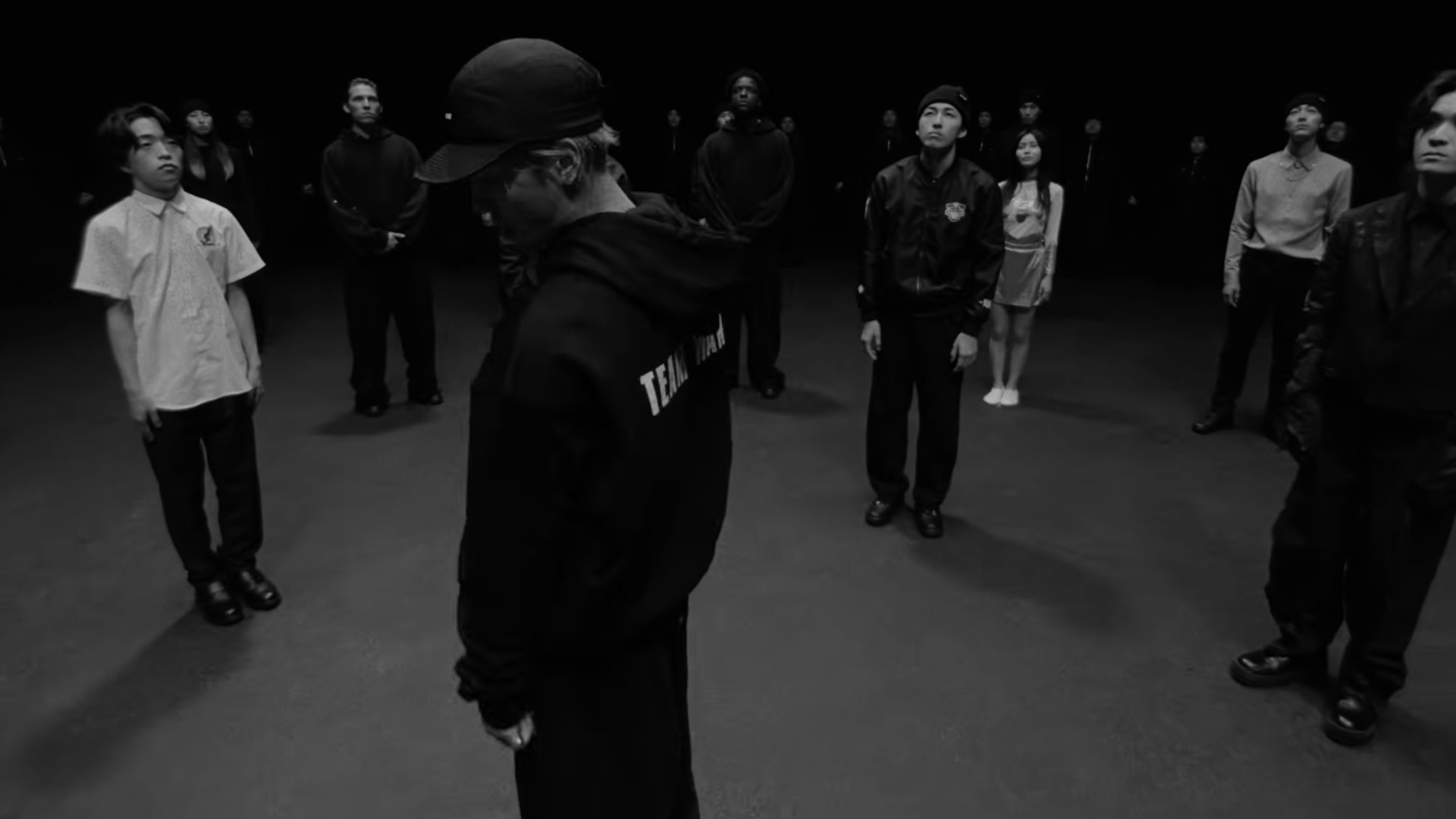 A group of people, including Jackson Wang, stand in a dimly lit space, each facing different directions. The image is in black and white.