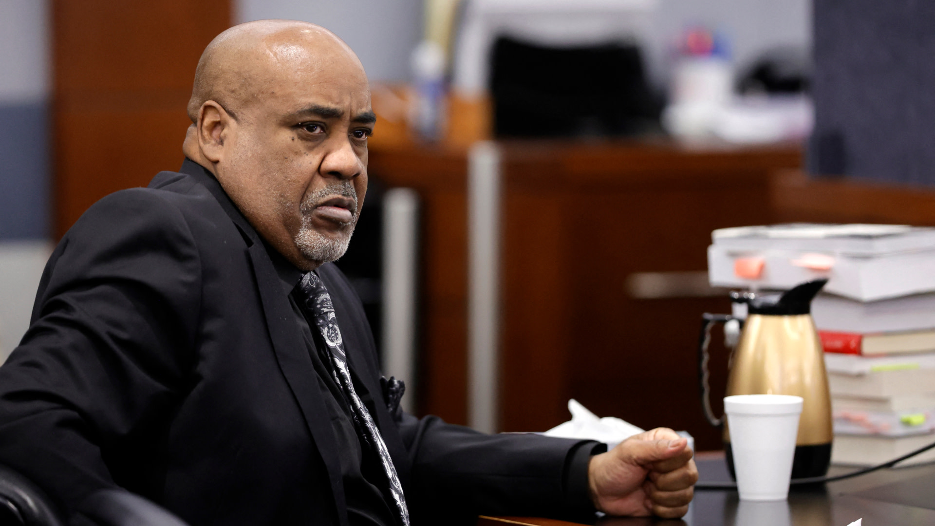 Duane "Keffe D" Davis in a suit sits at a table in a courtroom, looking serious. There are documents and a coffee pot on the table.