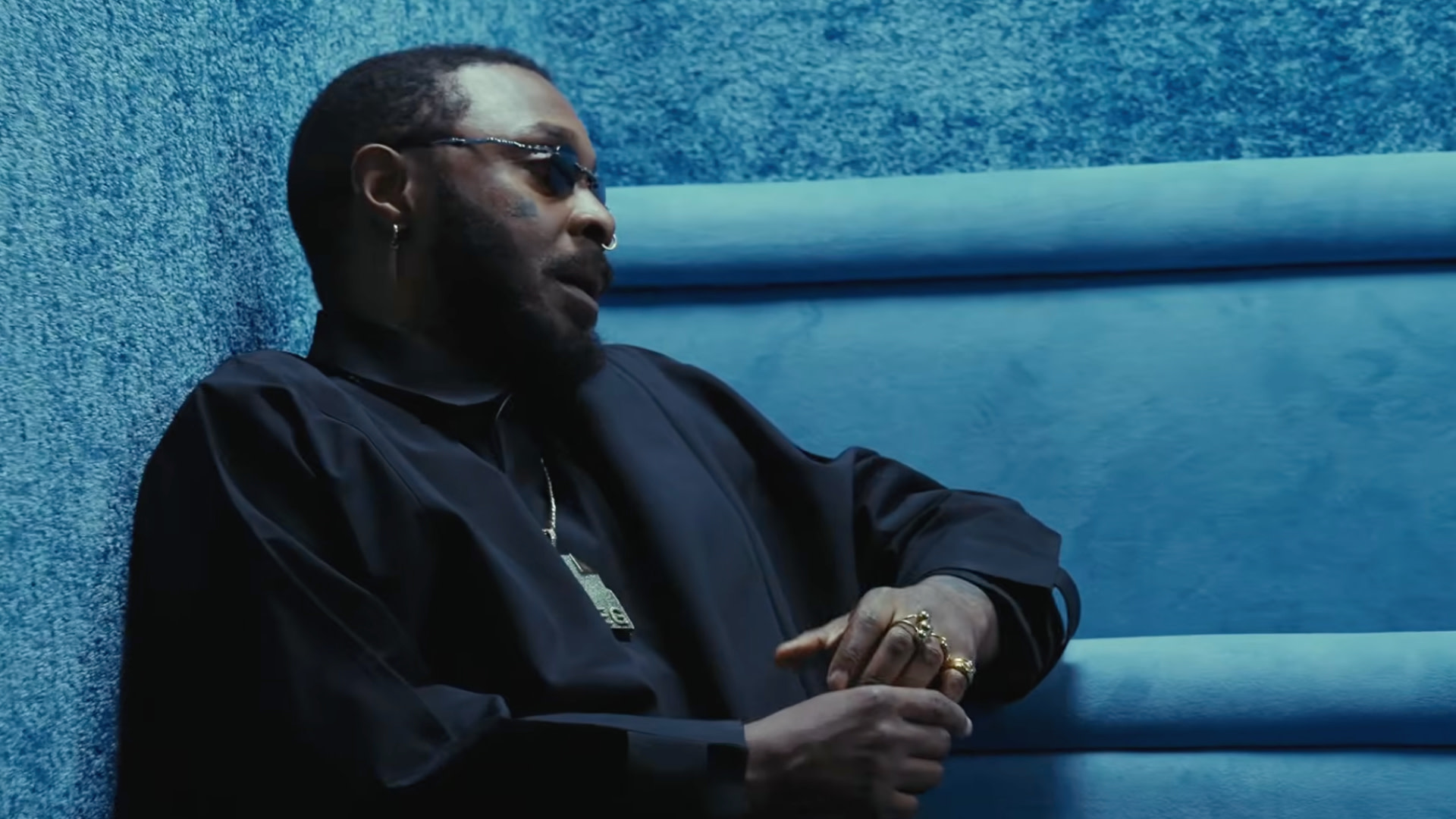 JPEGMAFIA with sunglasses and jewelry leans against a blue wall, wearing a black outfit.