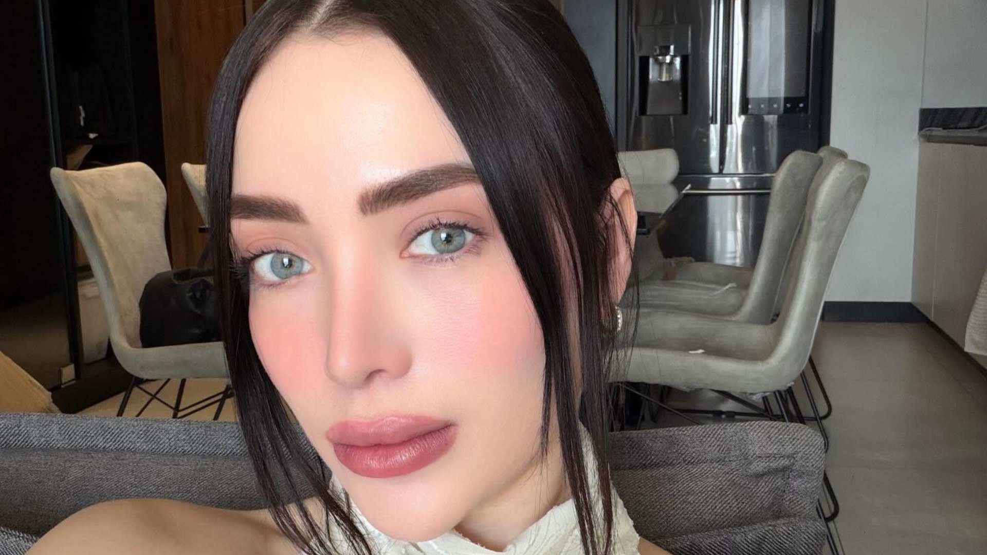 Influencer Adriana Garcia with dark hair and light eyes poses indoors, with a modern kitchen and dining area in the background.