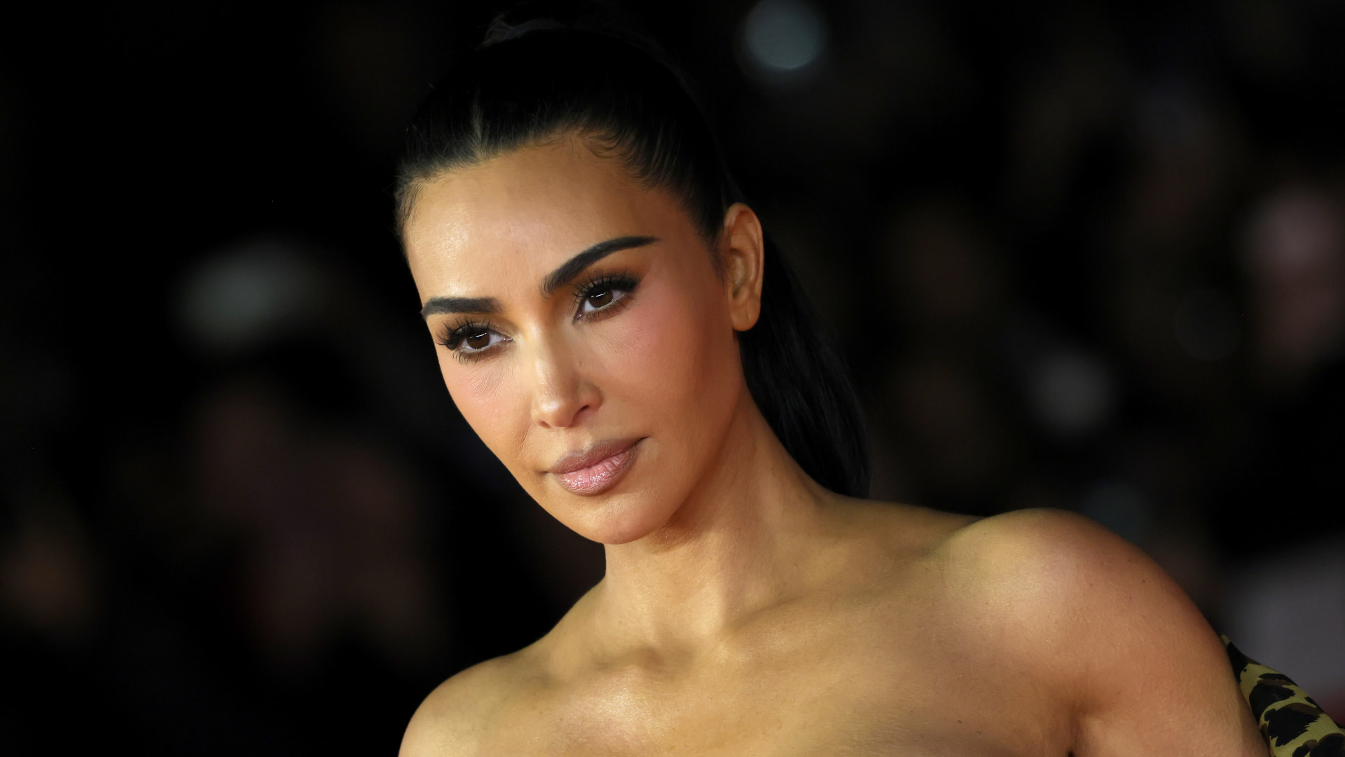 Kim Kardashian at an event, wearing a strapless outfit with sleek hair and subtle makeup.