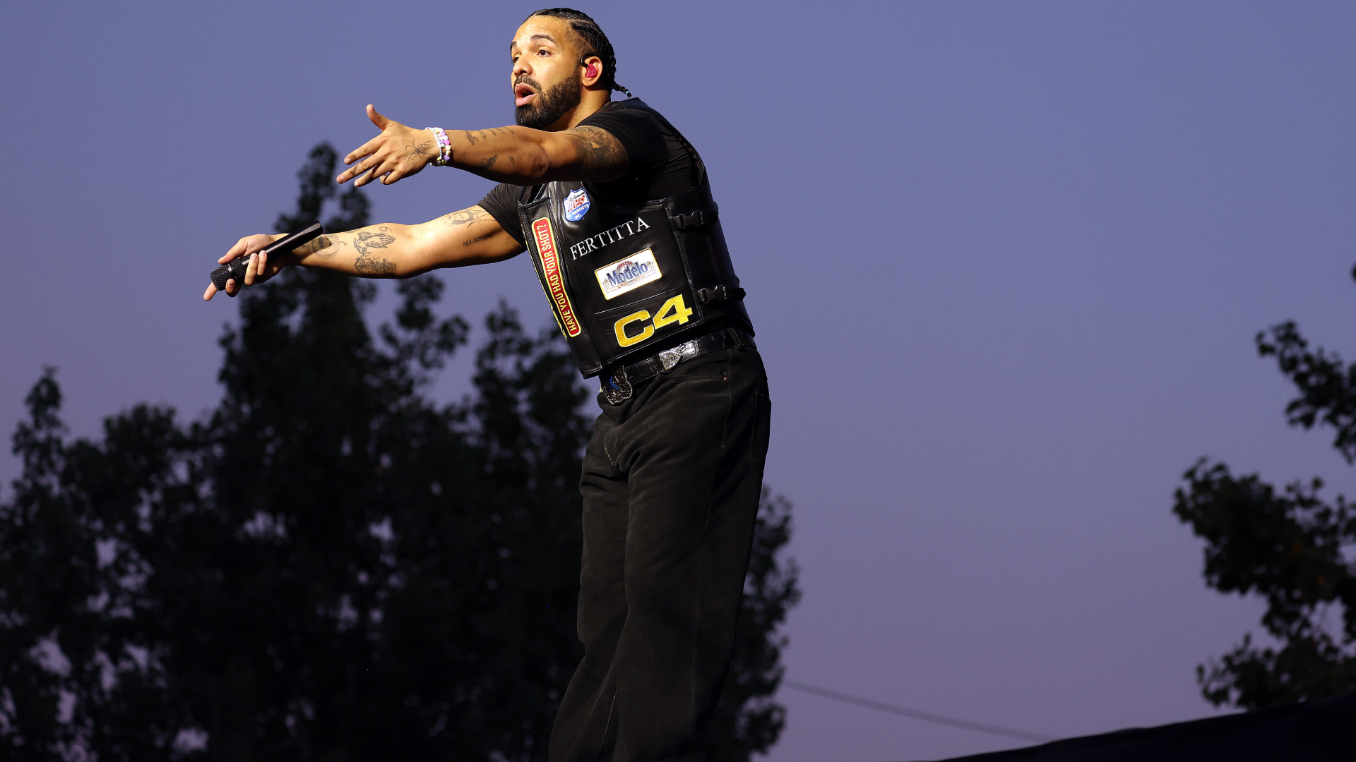 Drake performing on stage, wearing a black outfit with patches, gesturing passionately, against a twilight sky.