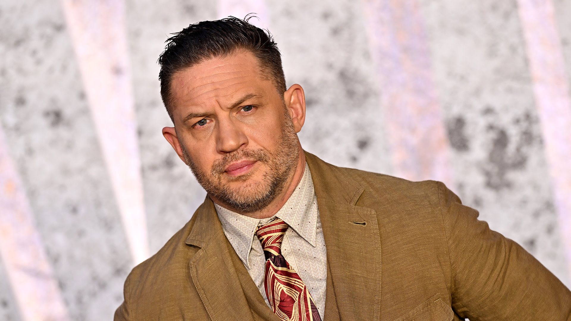 A man with short dark hair and facial hair, wearing a brown suit and patterned tie, poses against a textured background.