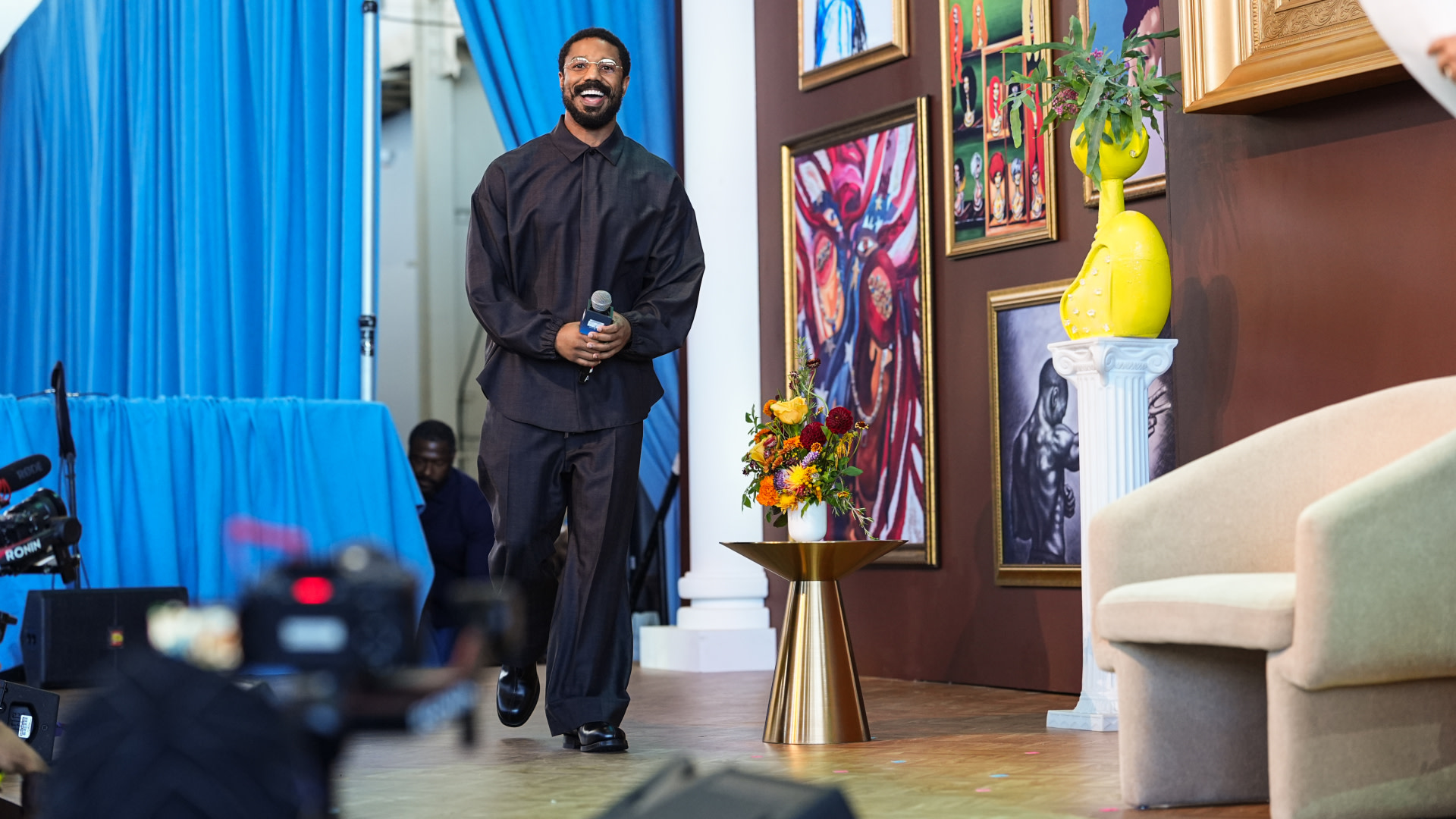 Michael B. Jordan in a dark outfit holding a microphone stands on a stage with colorful artwork and flowers. Blue curtains and a camera are visible.