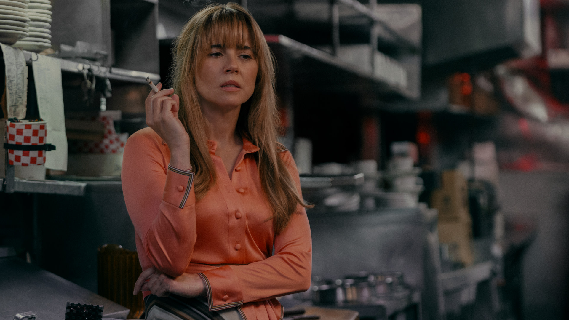 Linda Cardellini in a kitchen setting wearing an orange blouse, holding a cigarette, with a thoughtful expression.