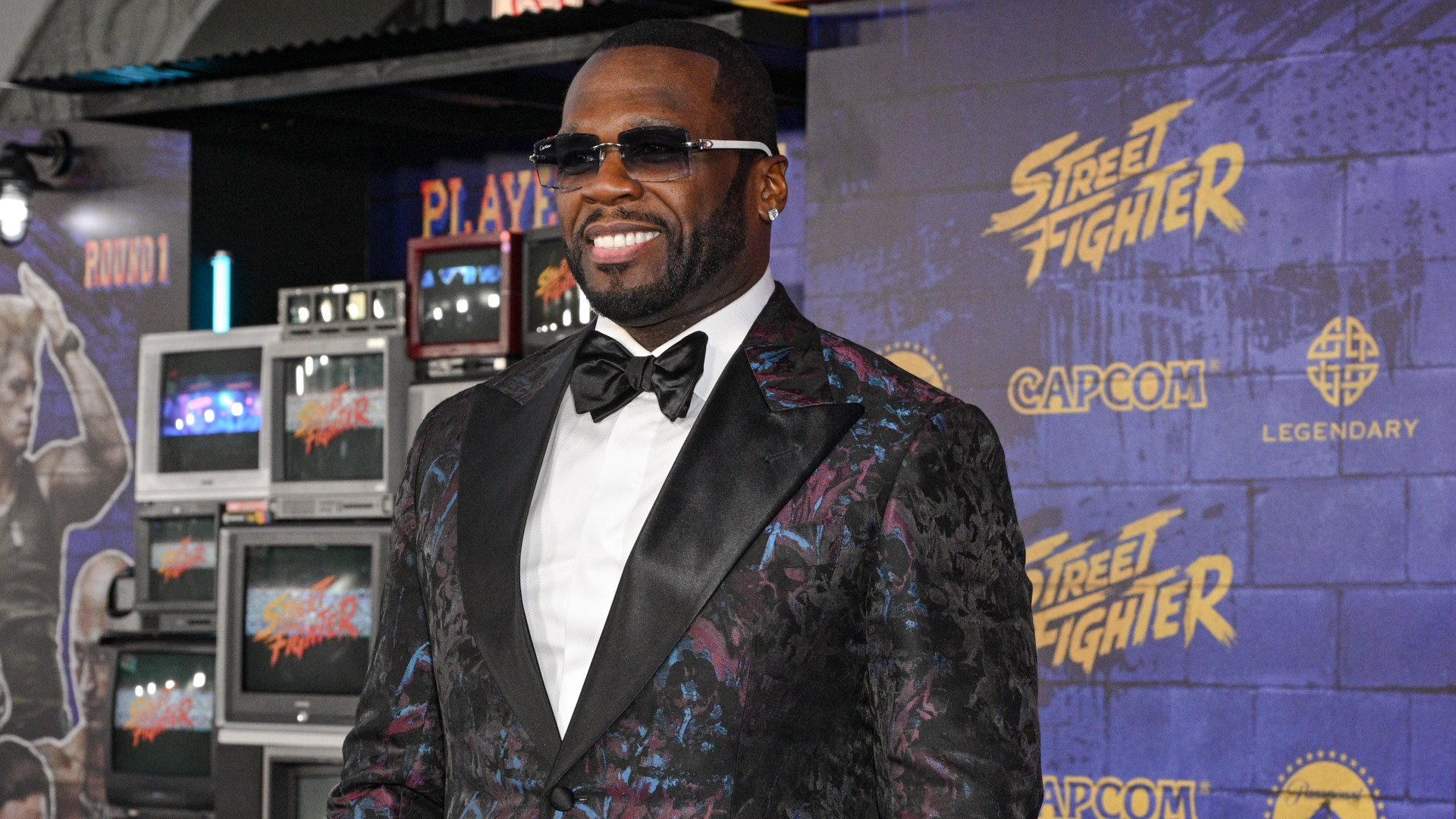 50 Cent in a floral suit and sunglasses smiles on the red carpet at a "Street Fighter" event with vintage TVs in the background.