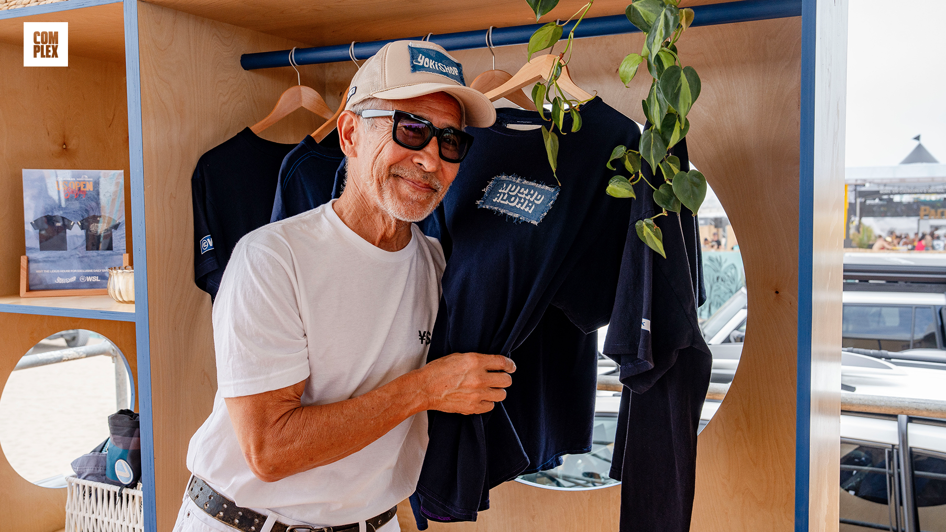 Man in sunglasses and a cap holding a navy blue shirt in a wooden booth, with plants hanging nearby.