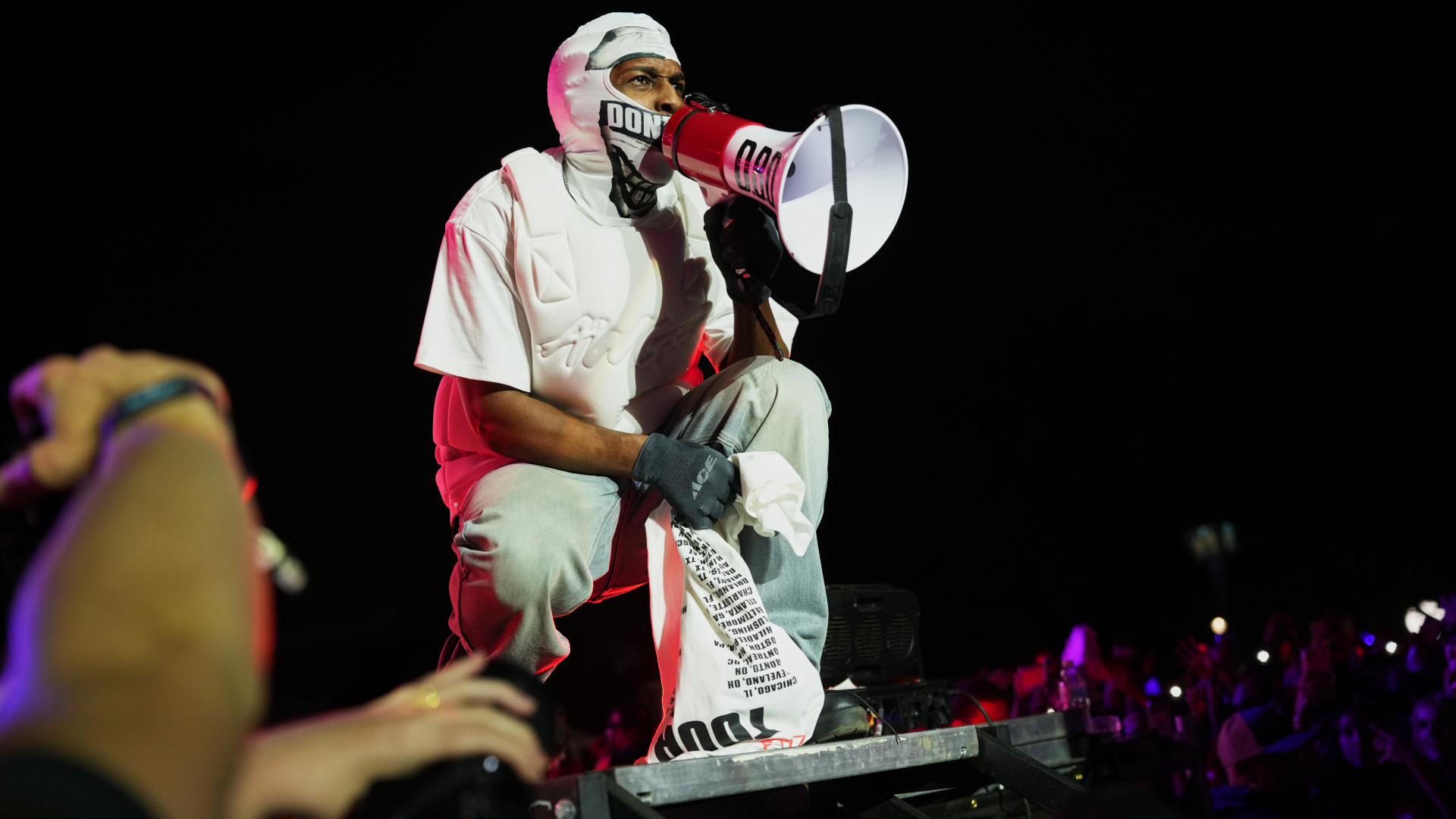 ASAP Rocky wearing a white outfit and mask, holding a megaphone on stage, with a crowd and colorful lights in the background.