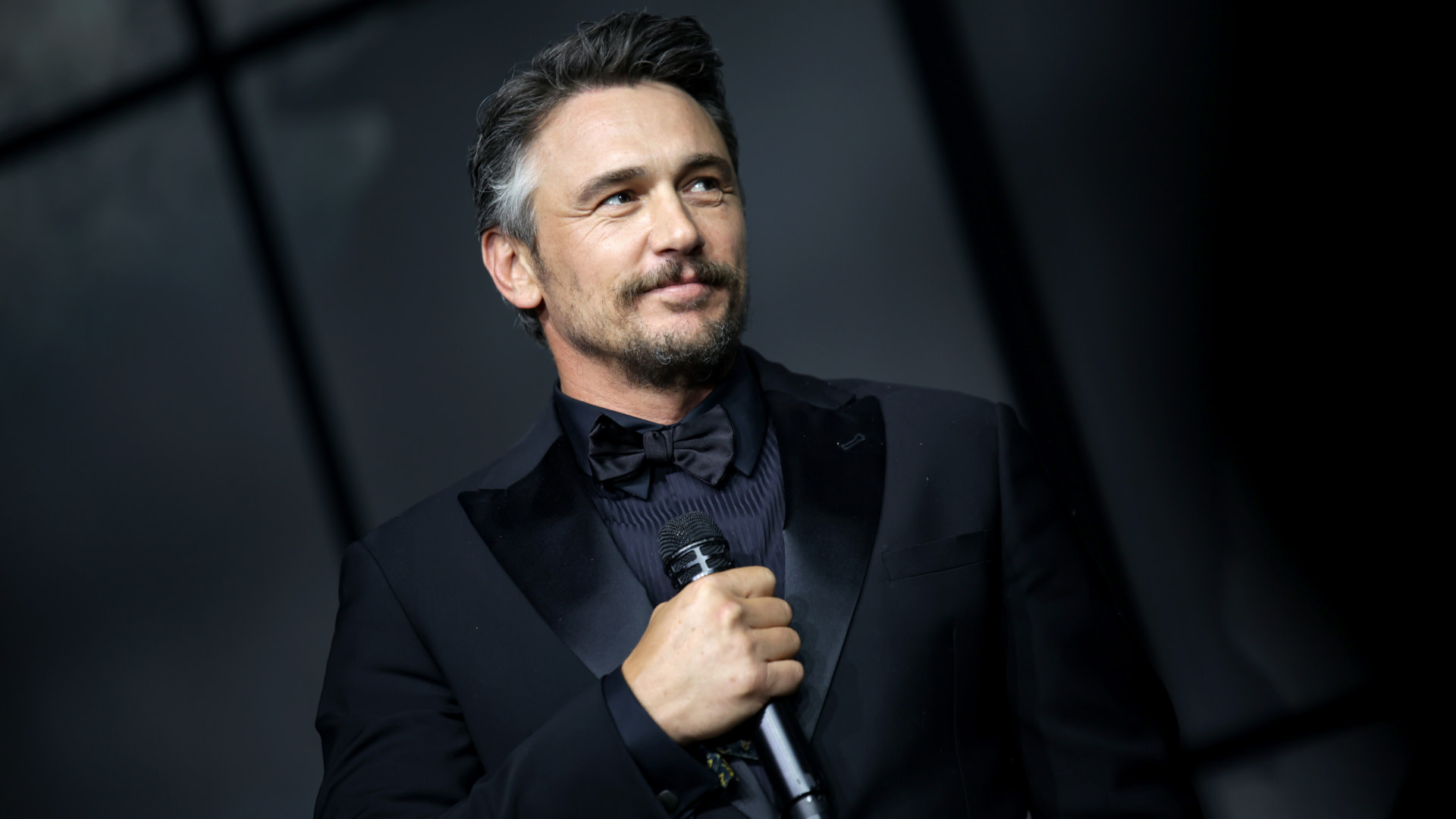 James Franco in a black tuxedo holds a microphone, standing against a dark backdrop.