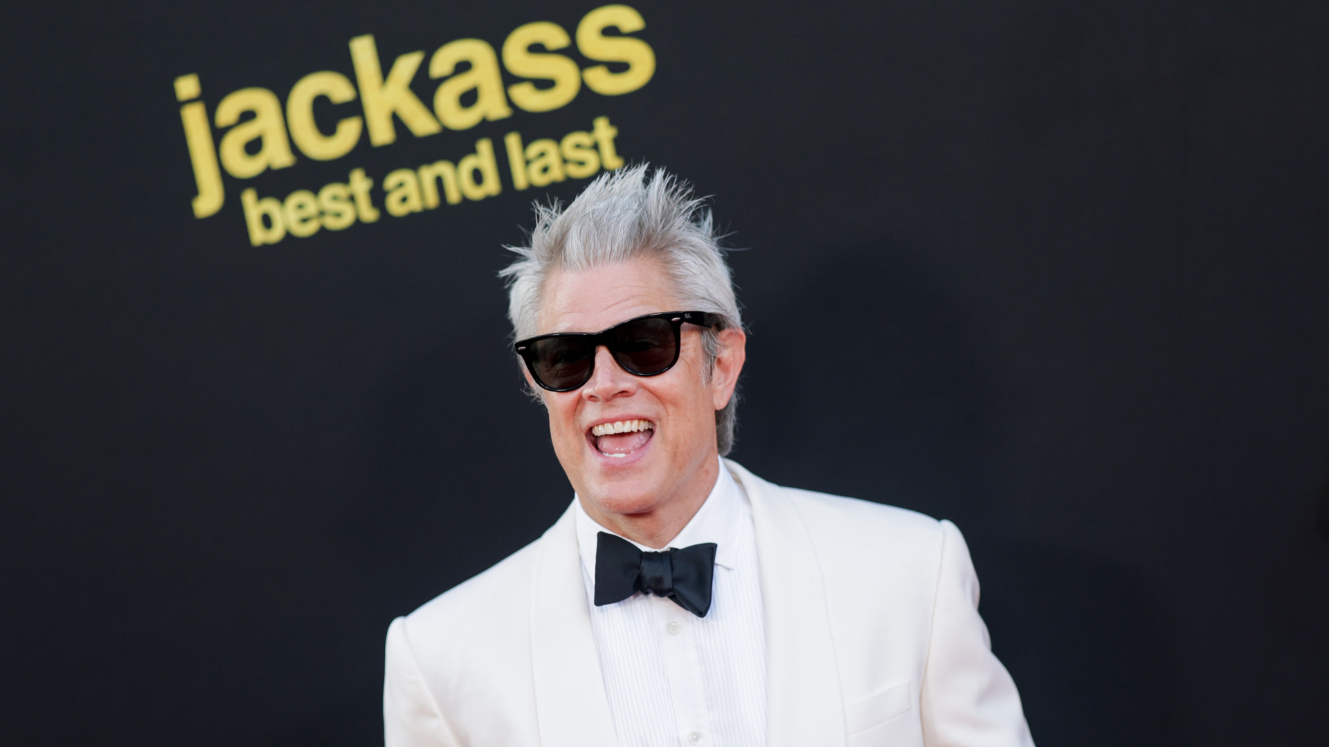 Johnny Knoxville in a white tuxedo and sunglasses smiles at a "Jackass" event backdrop.