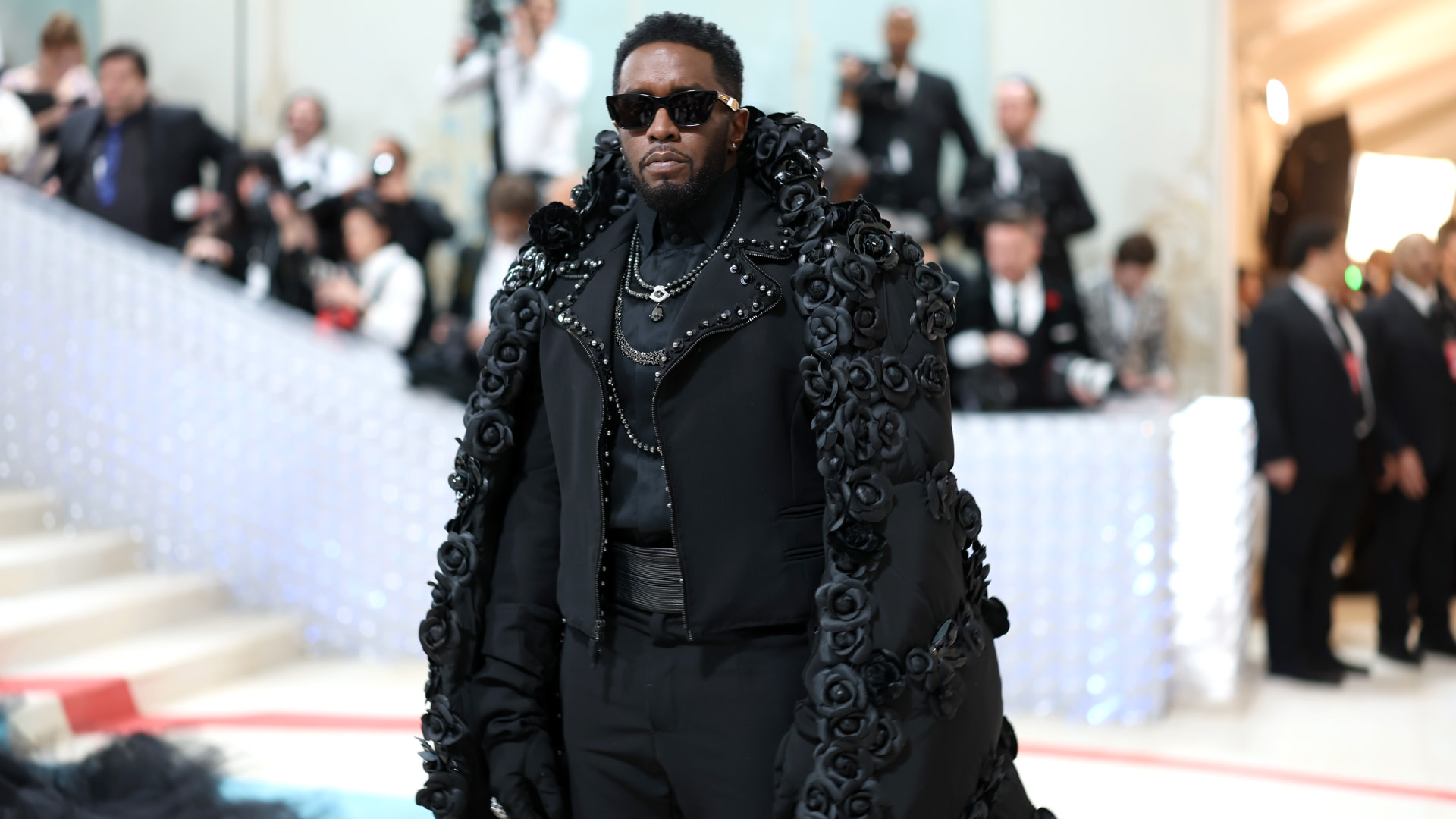 Diddy in a black, rose-embellished coat and sunglasses at a formal event, with photographers in the background.
