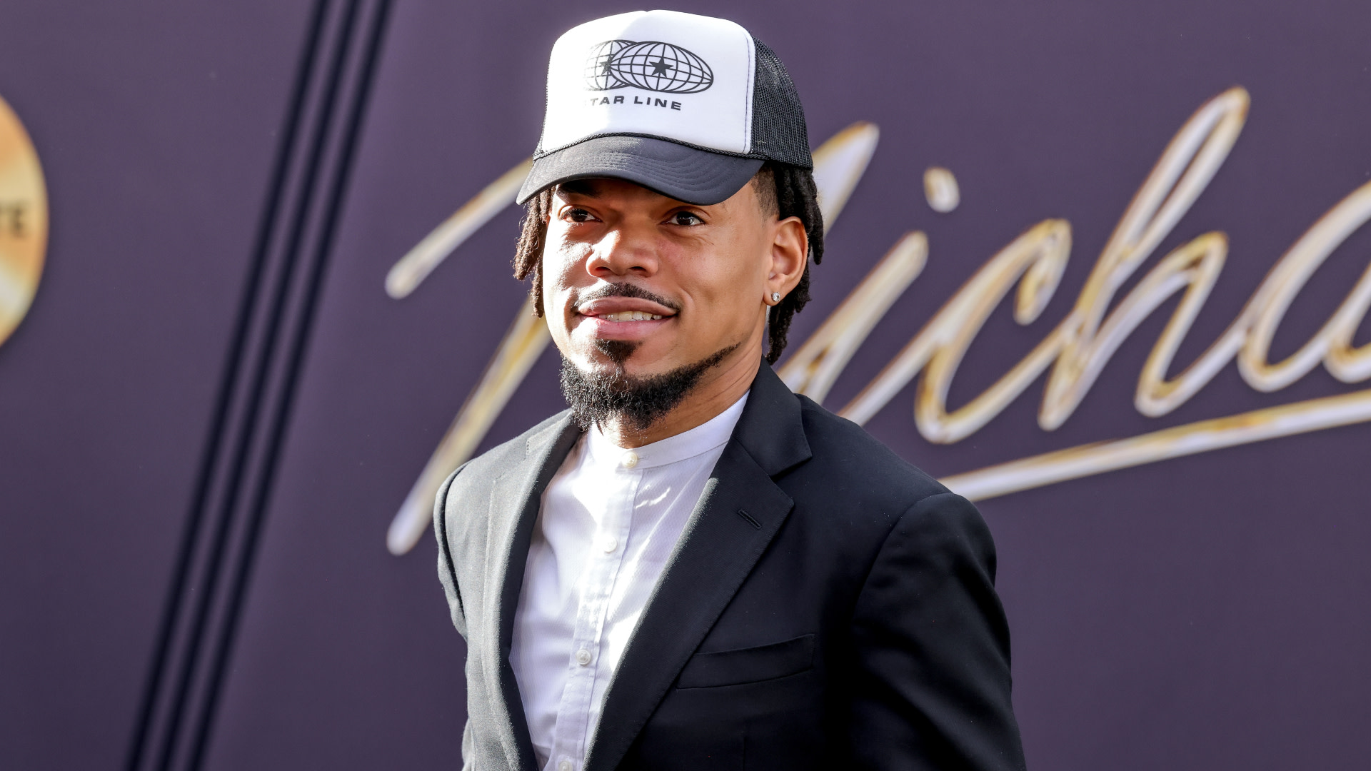 Chance the Rapper in a black suit and white shirt, wearing a black and white cap, smiles at an event.