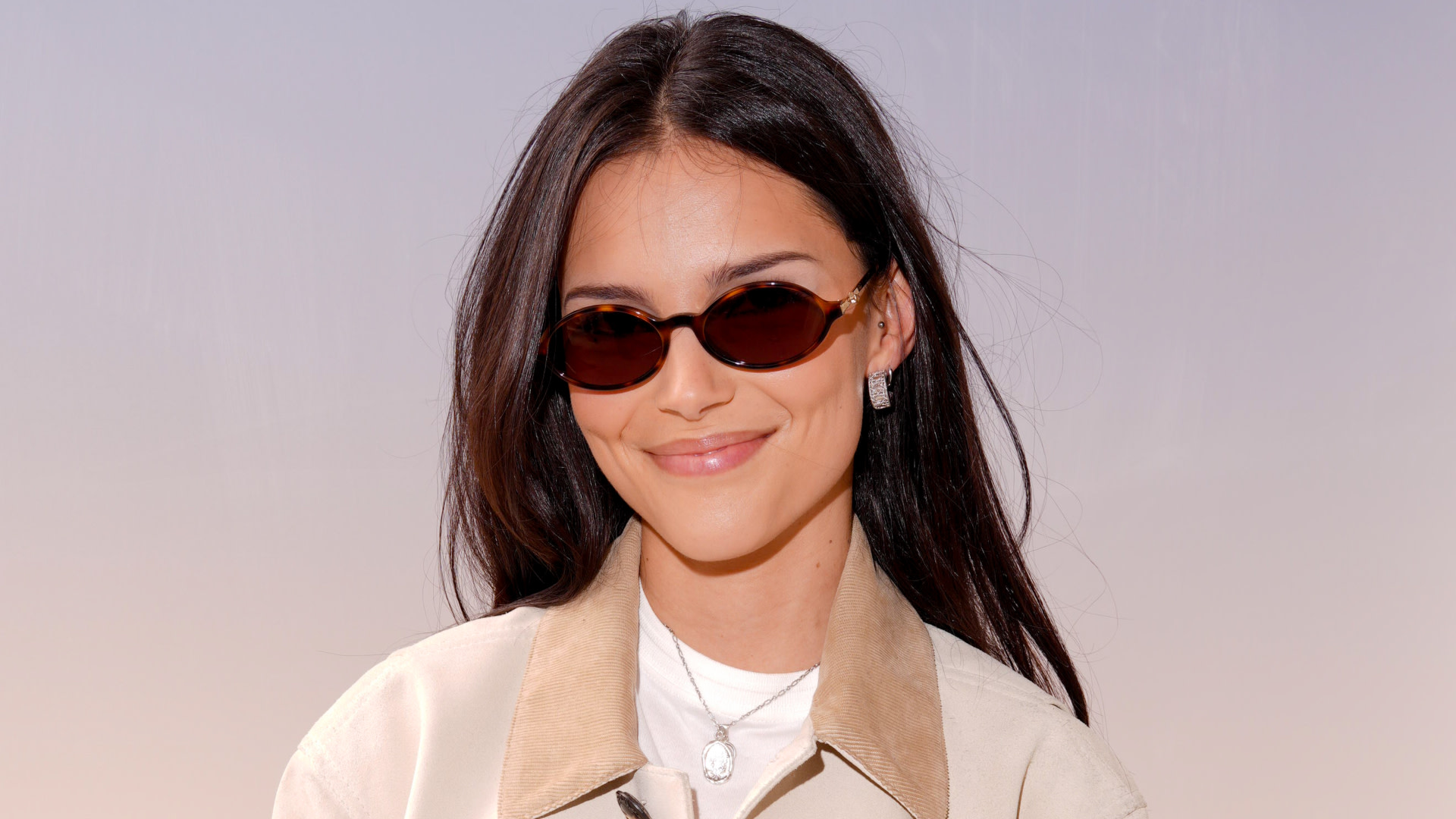 Inde Navarrette with long dark hair wearing sunglasses, earrings, and a light-colored jacket smiles at the camera.
