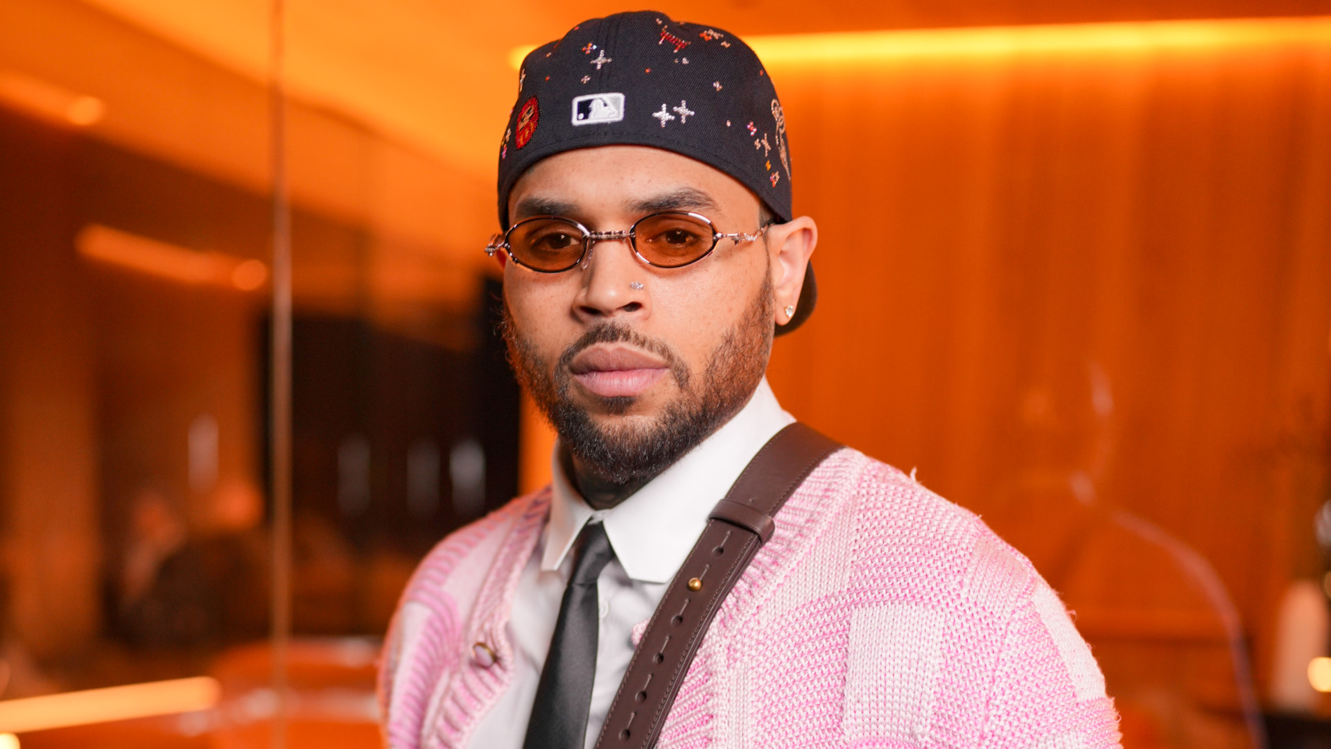 Chris Brown in a pink sweater, white shirt, and tie, wearing a black cap and sunglasses, stands in a warmly lit room.