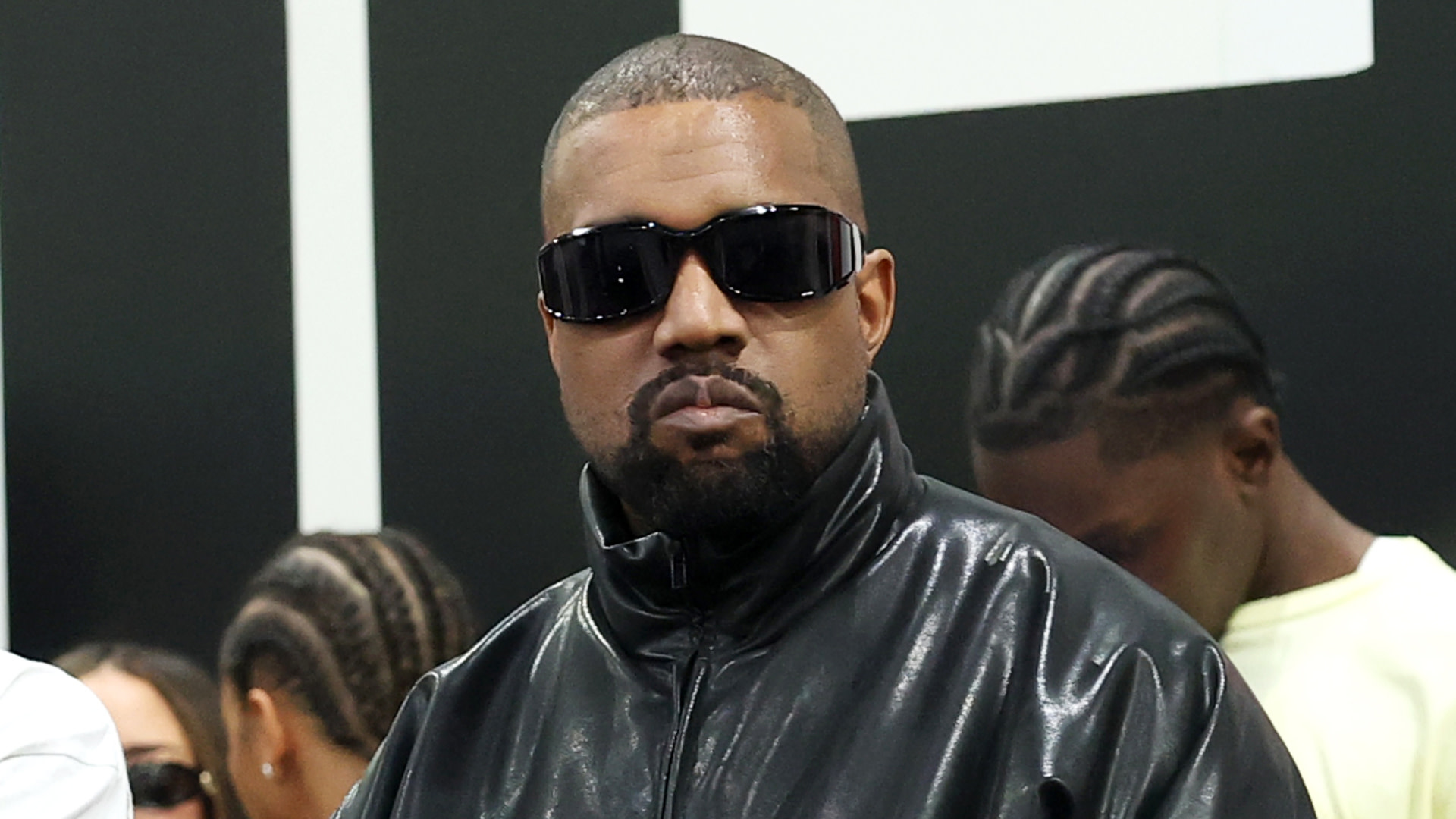 Kanye West wearing a black leather jacket and sunglasses, standing in front of a black and white background.