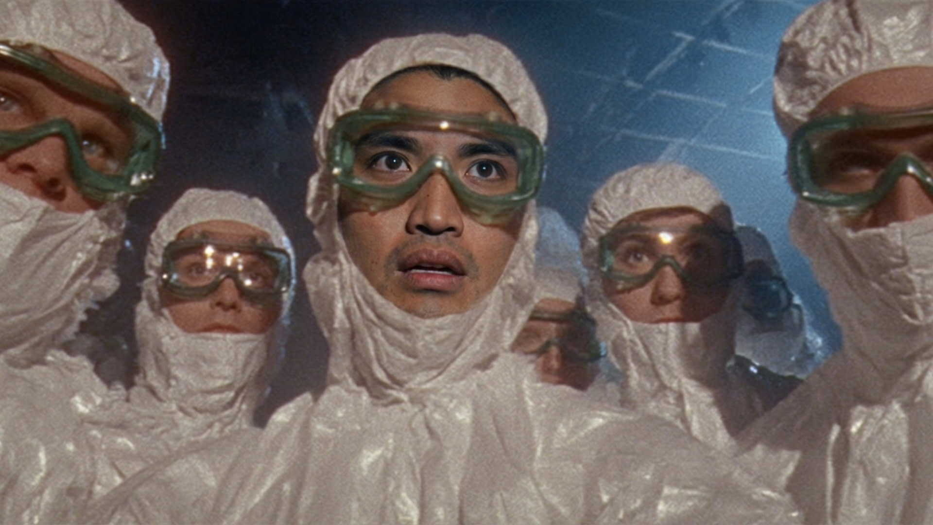 Chad Hugo and a group of people in white hazmat suits and green goggles, standing closely together, looking surprised or concerned.