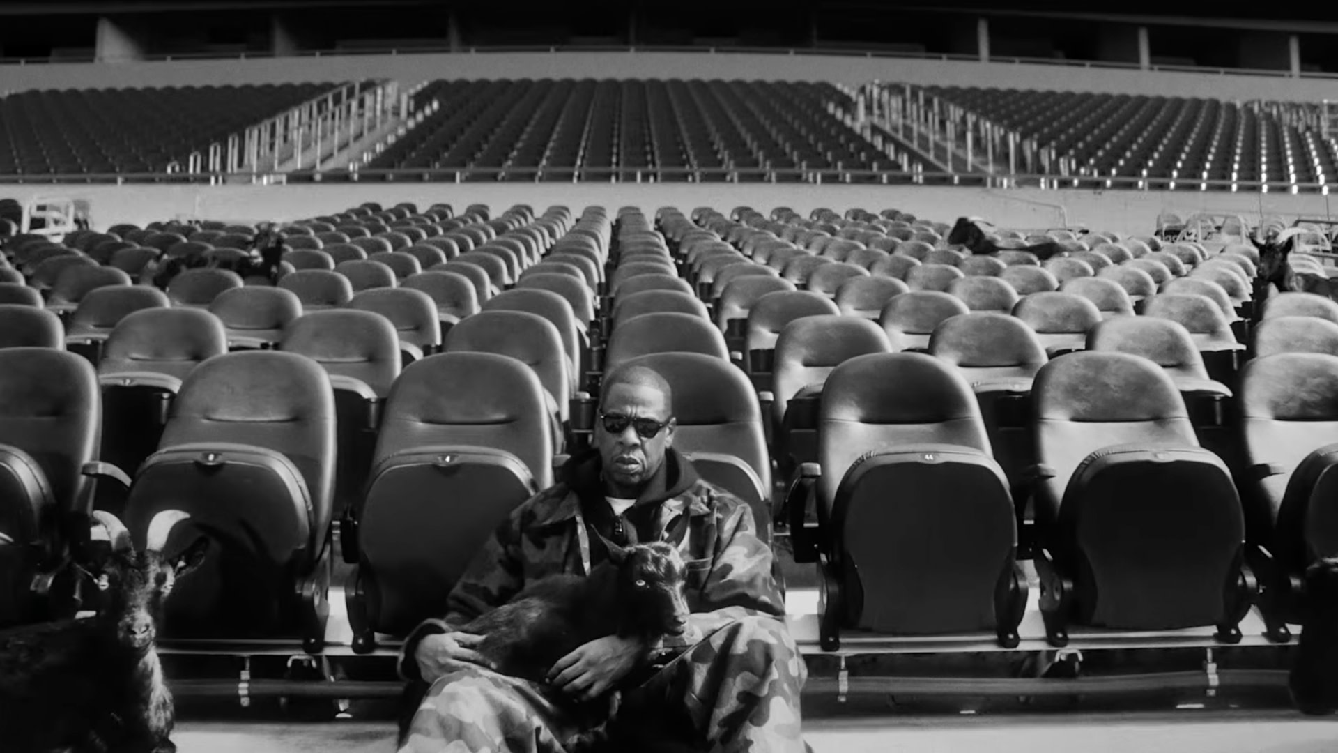 Jay-Z in camouflage sits in an empty stadium holding a goat, with several goats around him. The image is in black and white.